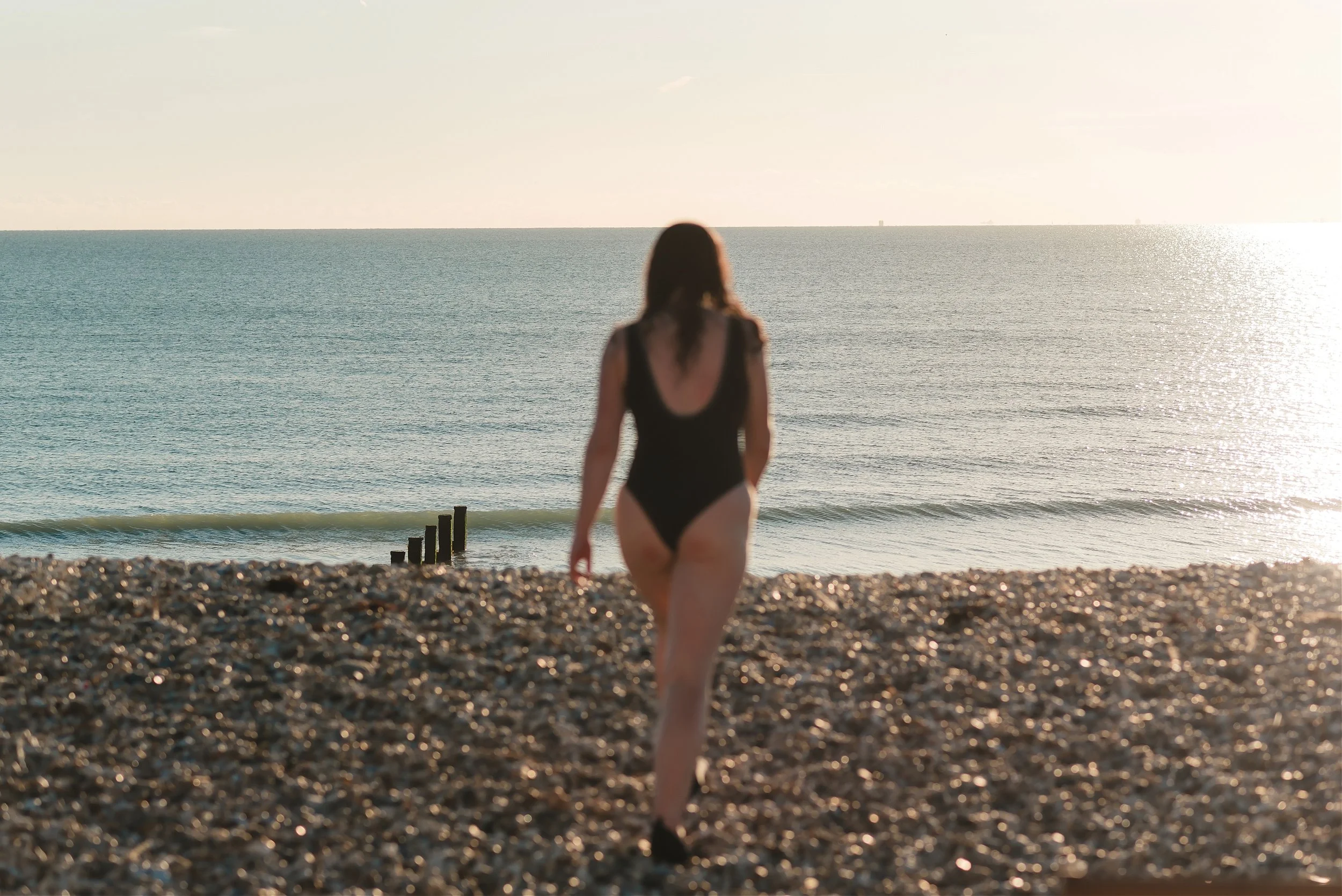 Woman in a black swimsuit walking across a shingle beach towards the sea for wild swimming in West Sussex