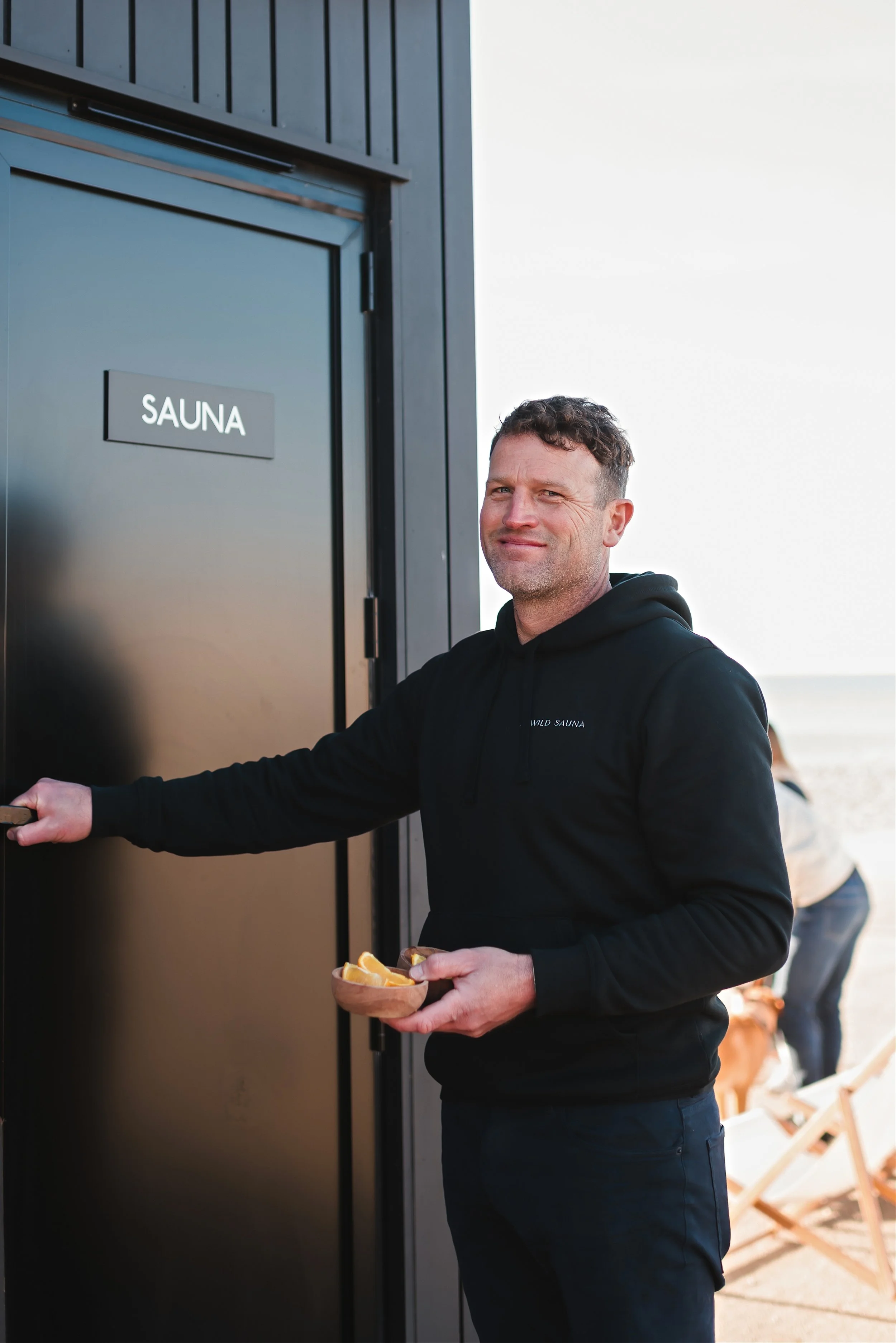 A man standing outdoors next to a black sauna door labeled 'SAUNA', holding a small bowl of food and smiling at the camera. The background shows a beach setting with dunes and a wooden chair.