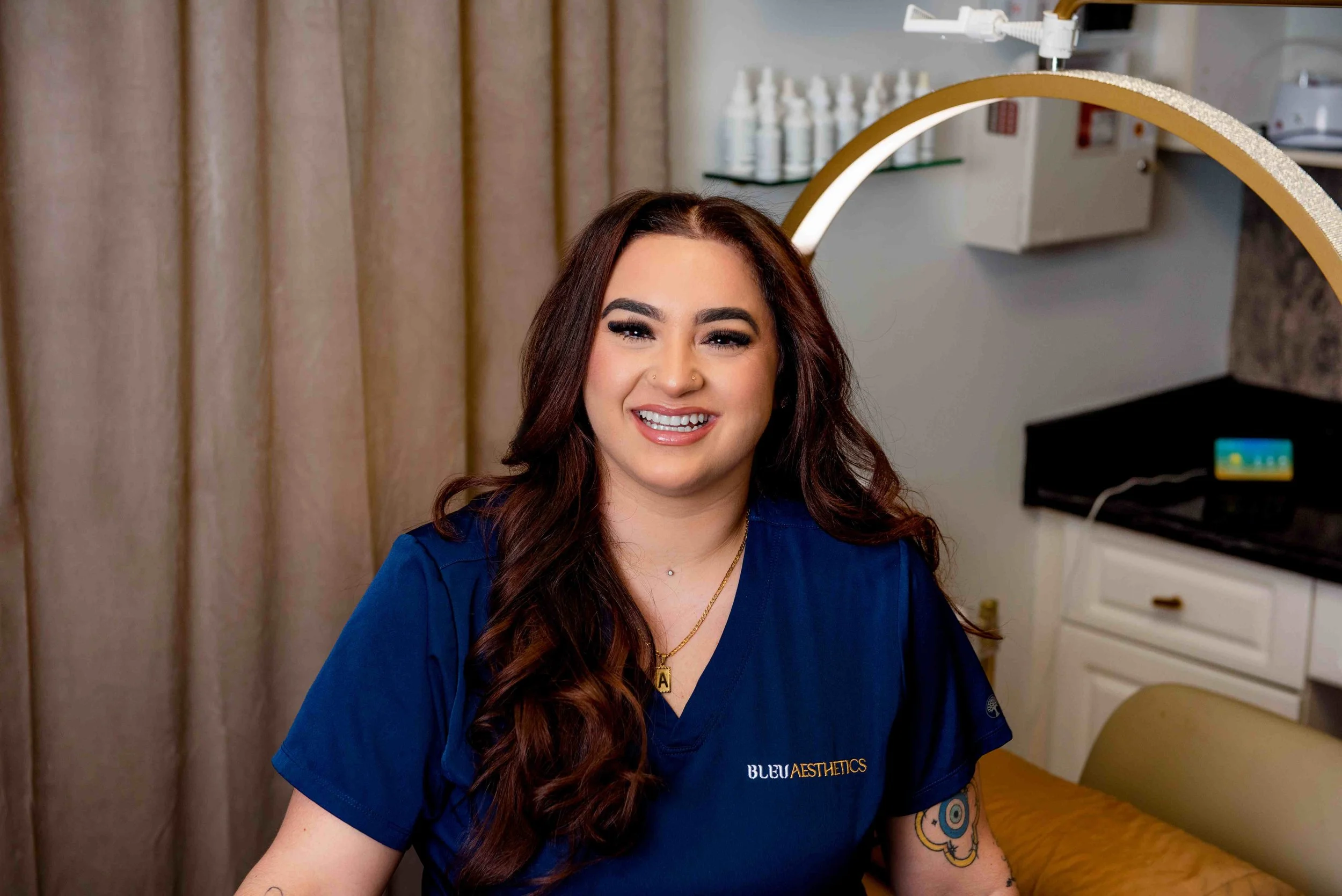 A smiling woman with long wavy brown hair wearing a blue medical uniform with a badge that says BULE AESTHETICS, sitting in a medical office or spa room.