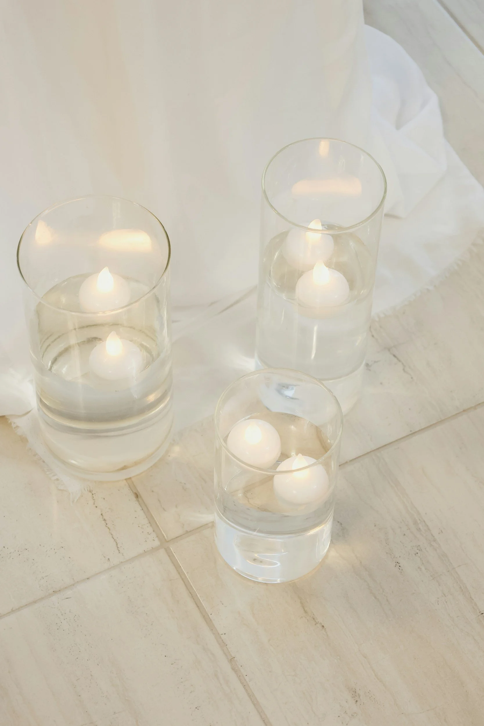 Multiple glass vases with floating candles on a marble surface, white tablecloth in the background.