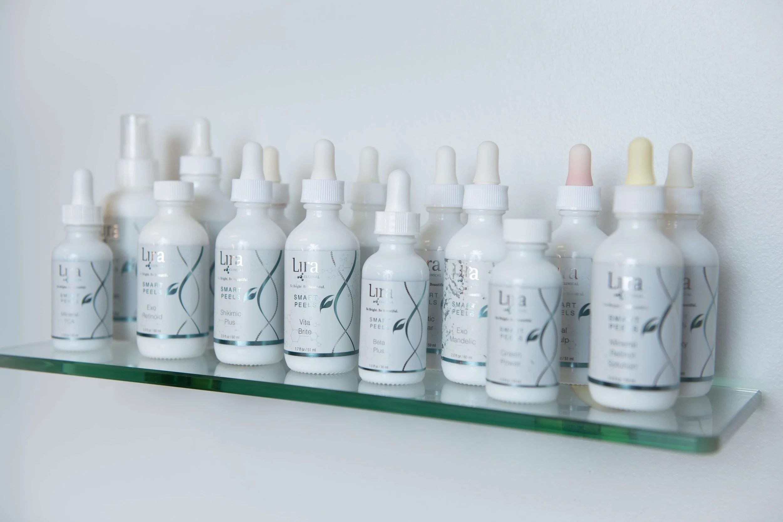 A row of white skincare bottles with dropper caps displayed on a glass shelf against a white wall.