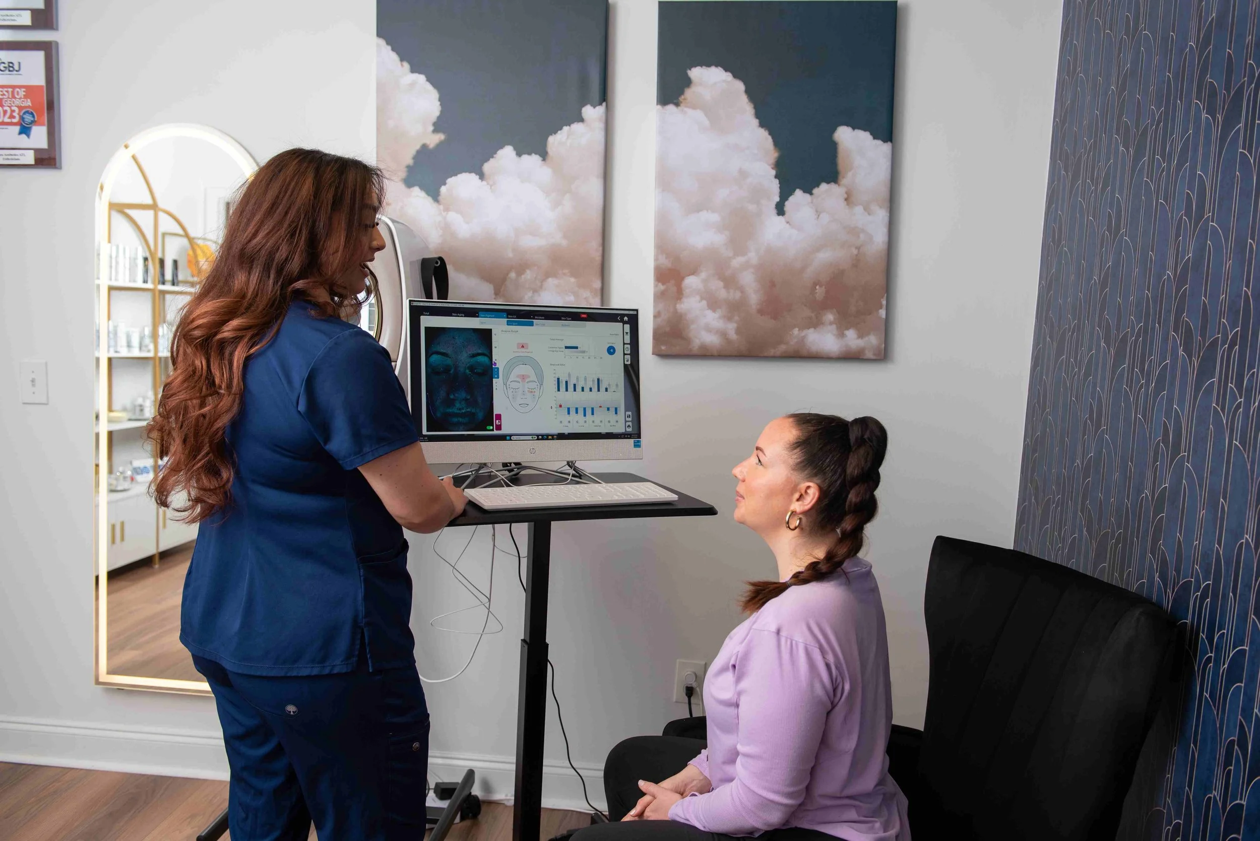A healthcare professional with long, wavy red hair wearing blue scrubs talking to a seated woman with dark hair in braids, wearing a light purple shirt, inside a modern medical office with cloud-themed artwork on the wall and a computer displaying facial analysis data.