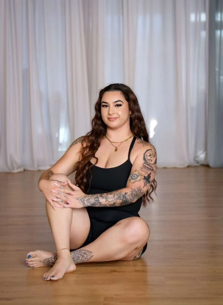 A woman sitting on a wooden floor with white curtains in the background. She has long, wavy brown hair, tattoos on her arms and leg, and is wearing a black tank top and shorts.
