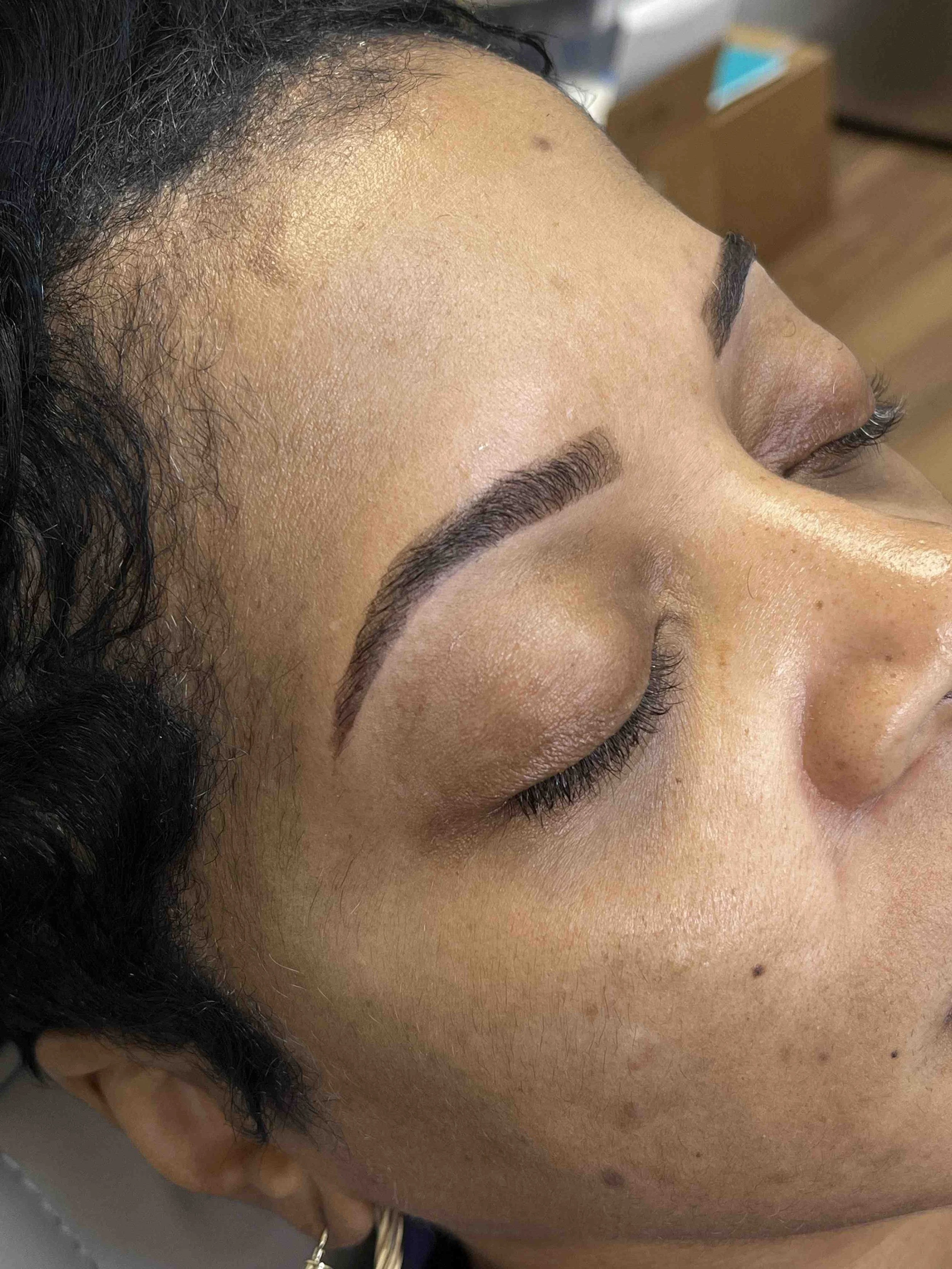 A close-up view of a woman's face and eyebrow, with her eyes closed, showing clear skin, even complexion, and professionally shaped eyebrows.