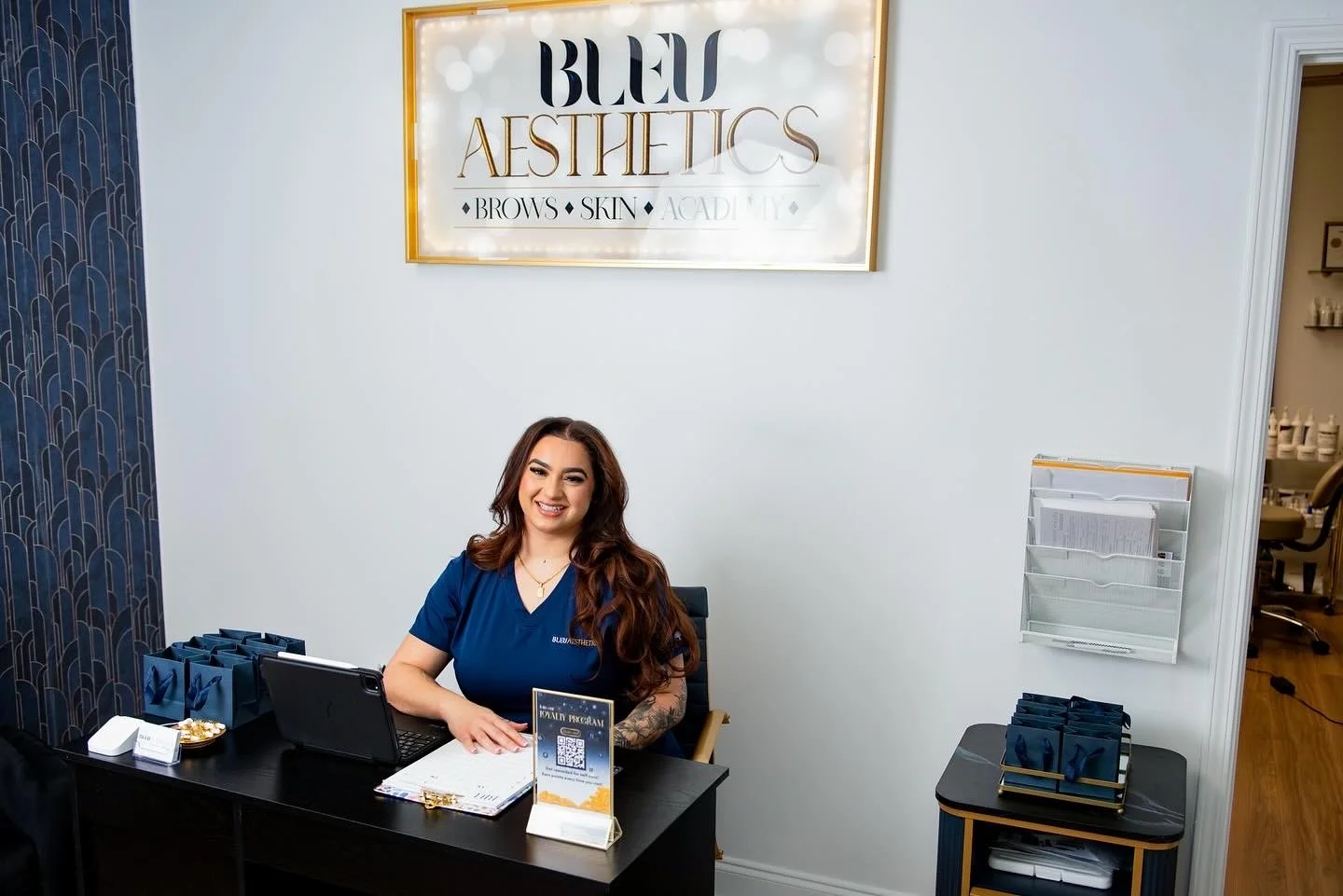 A woman with long brown hair, wearing a blue scrubs, smiling at the reception desk of an aesthetics clinic named Blue Aesthetics. A sign with the clinic's name is on the wall behind her. The desk has a laptop, a sign with a QR code, and blue gift bags. There are file holders and pamphlets on the right side of the wall, with a doorway leading to another room in the background.