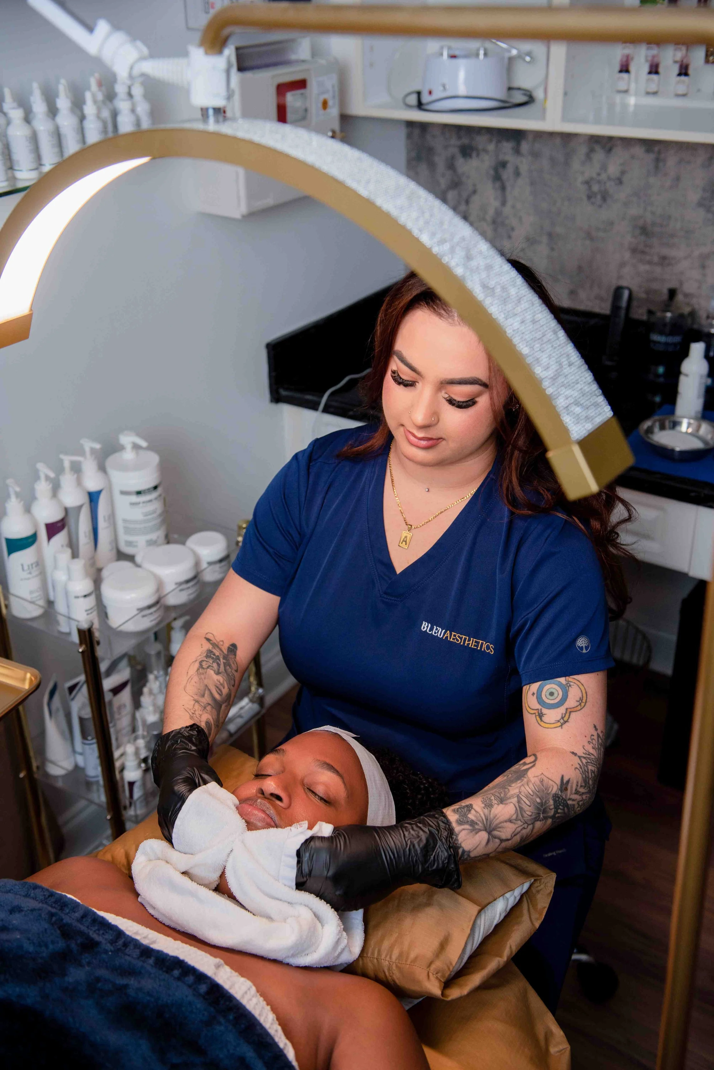 Aesthetician performing a facial treatment on a woman lying on a treatment bed in a spa or skincare clinic, with skincare products on shelves in the background.