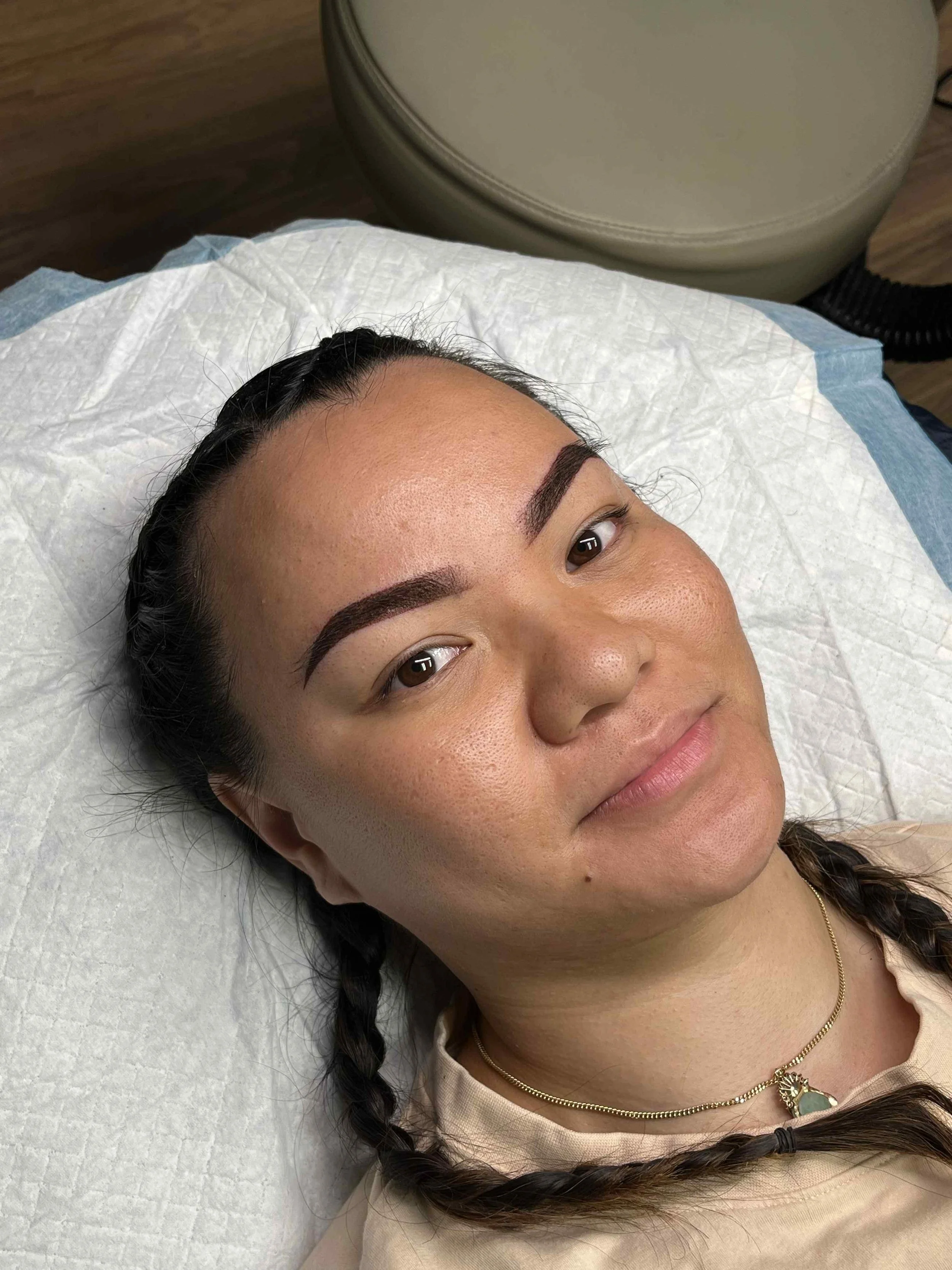 A woman lying on a white medical sheet, showing her face with filled-in eyebrows and braided hair, with a beige chair and wooden floor in the background.