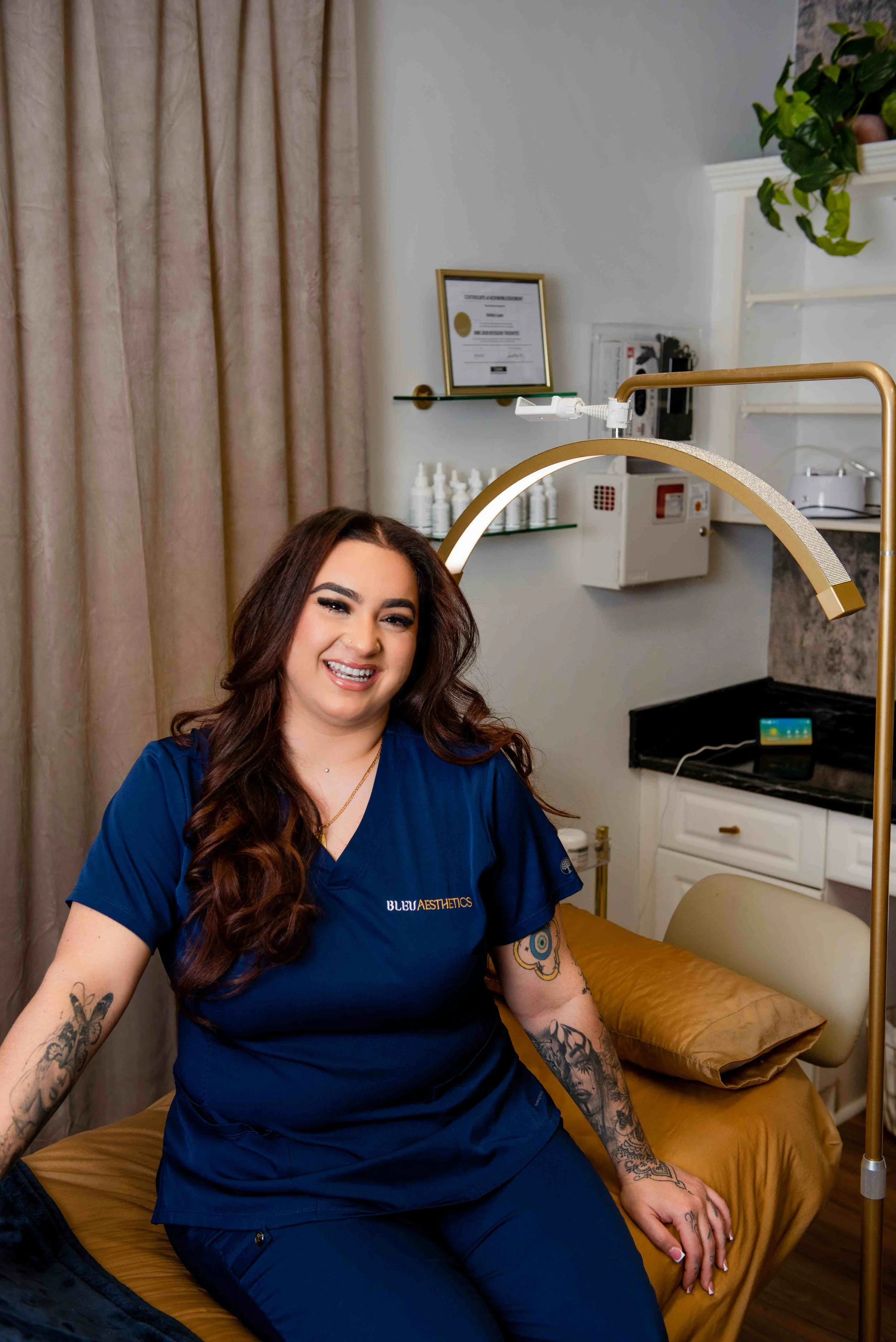 A woman with long brown hair and tattoos on her arms, dressed in blue medical scrubs, smiling and sitting on a medical examination table covered with a brown sheet in a clinic room.