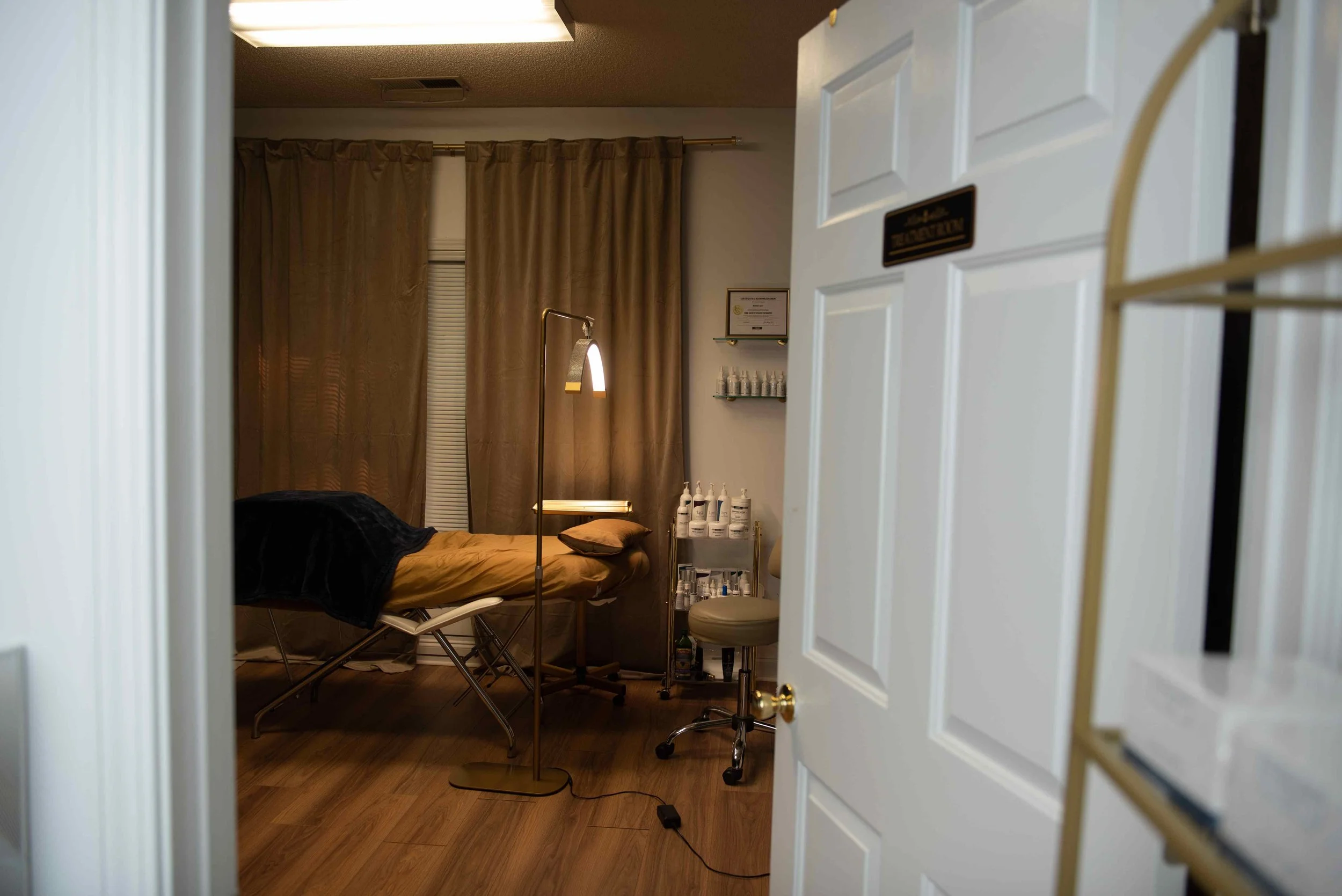 View of a room with a massage or examination table, a floor lamp, shelves with bottles, and closed curtains, seen through a door.