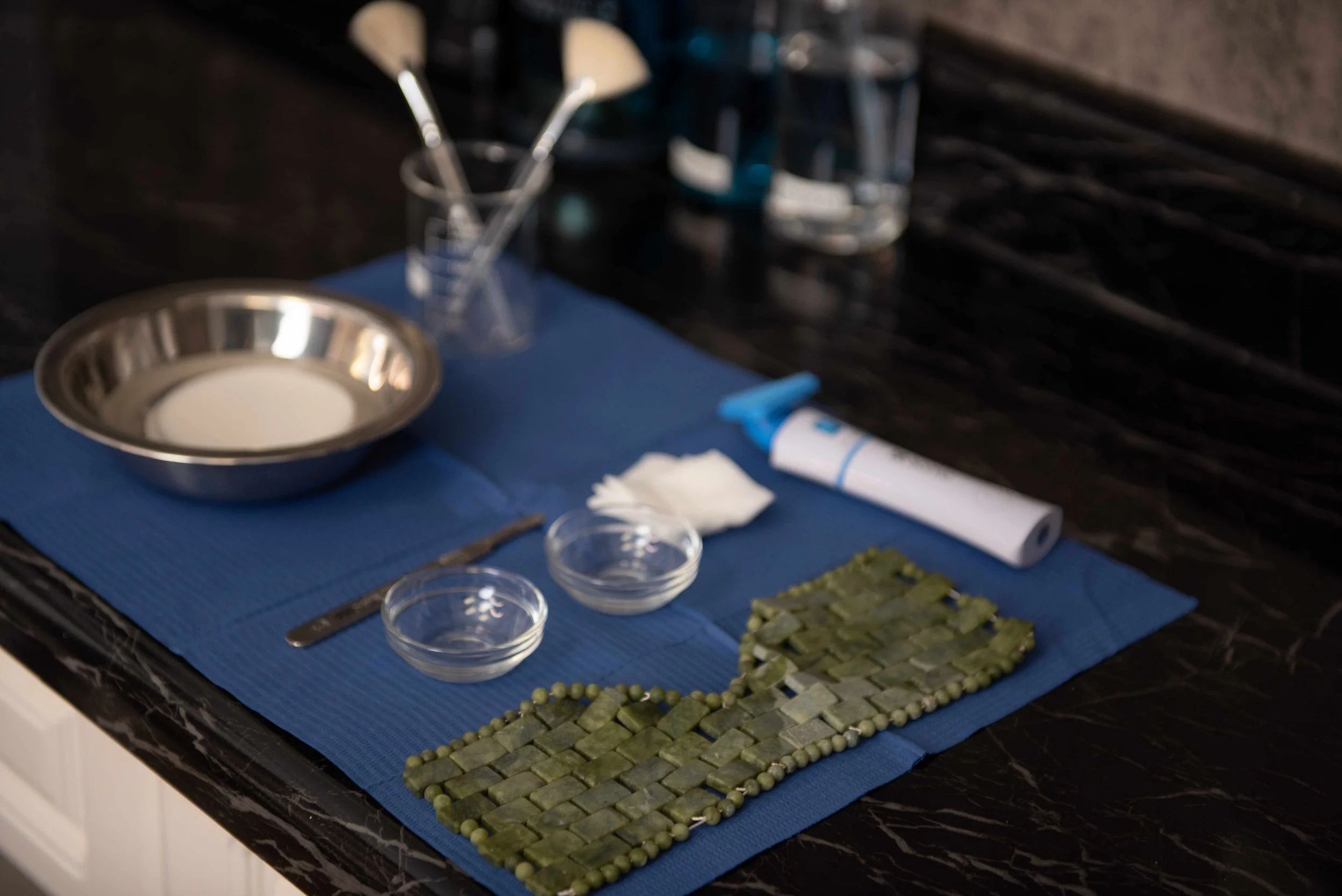 A kitchen countertop with a blue mat displaying stones, a metal bowl, glass jars, small bowls, and some utensils, with a black textured backsplash.