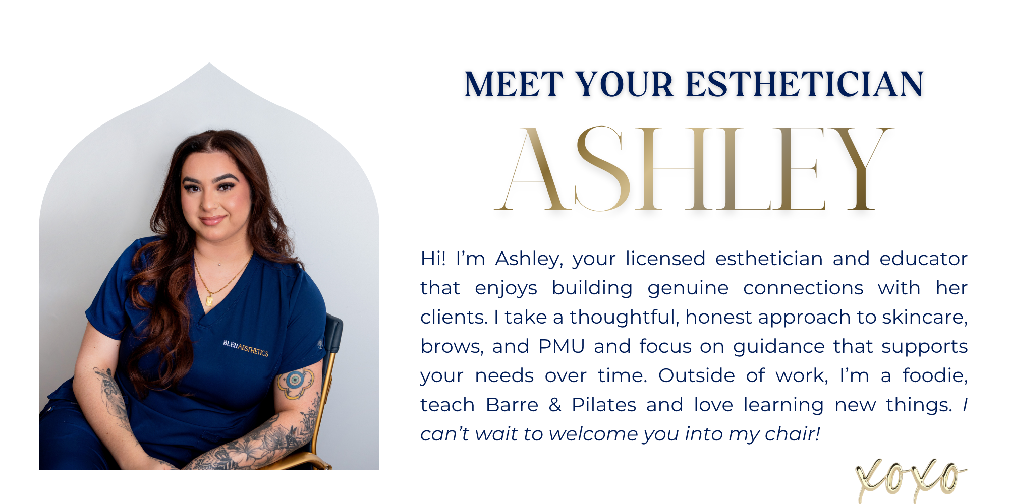 A woman with long brown hair, tattoos on her arms, wearing blue scrubs, smiles at camera, sitting in front of a white wall.