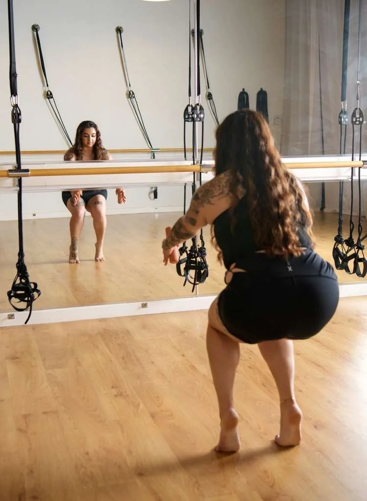 A woman with long dark hair in a black outfit practices a fitness squat in front of a mirror, in a fitness studio with wooden floors and resistance bands hanging on the wall.