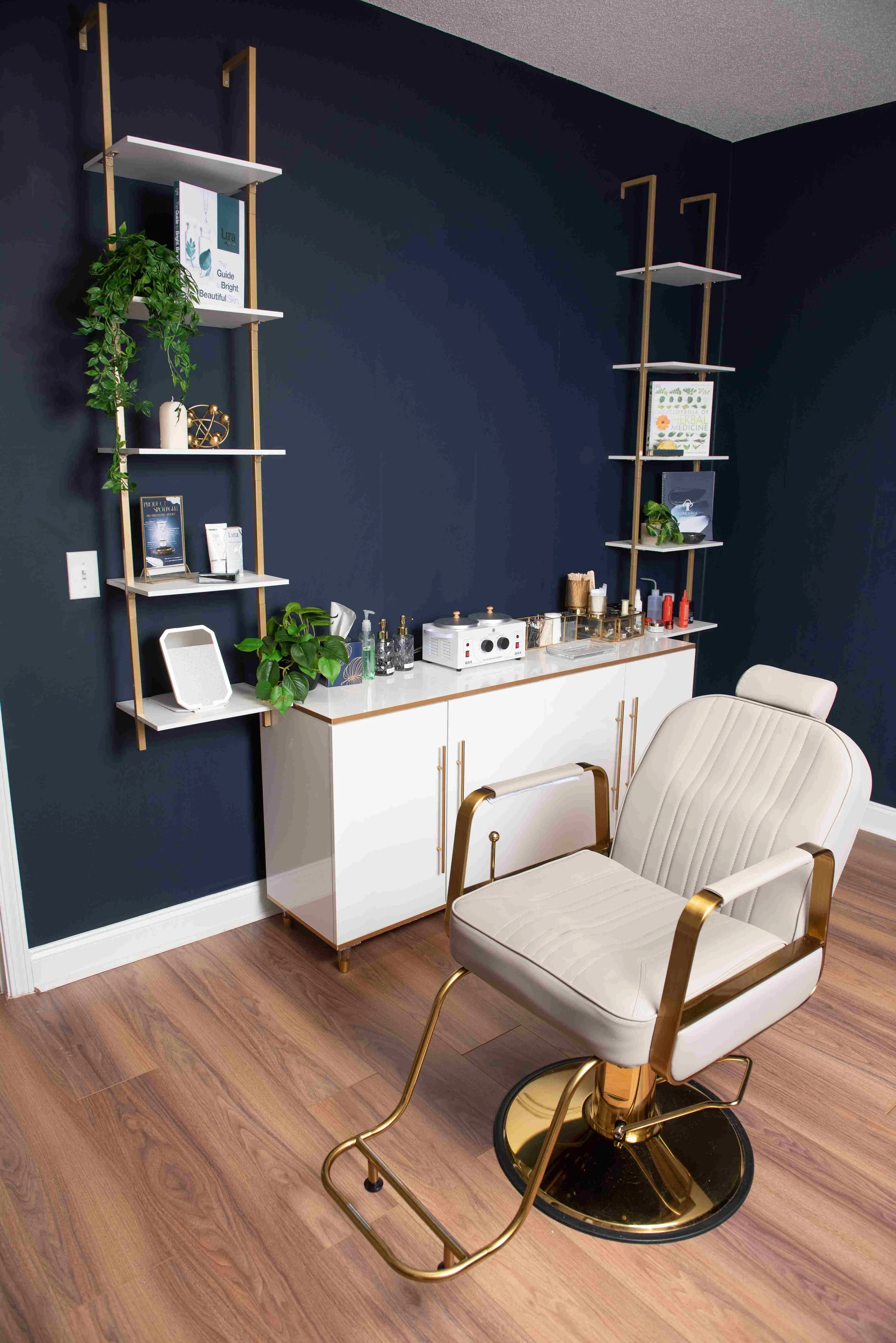 Interior of a room with a white and gold office chair, dark blue walls with white and gold shelving units, a white cabinet with various supplies, and wooden flooring.