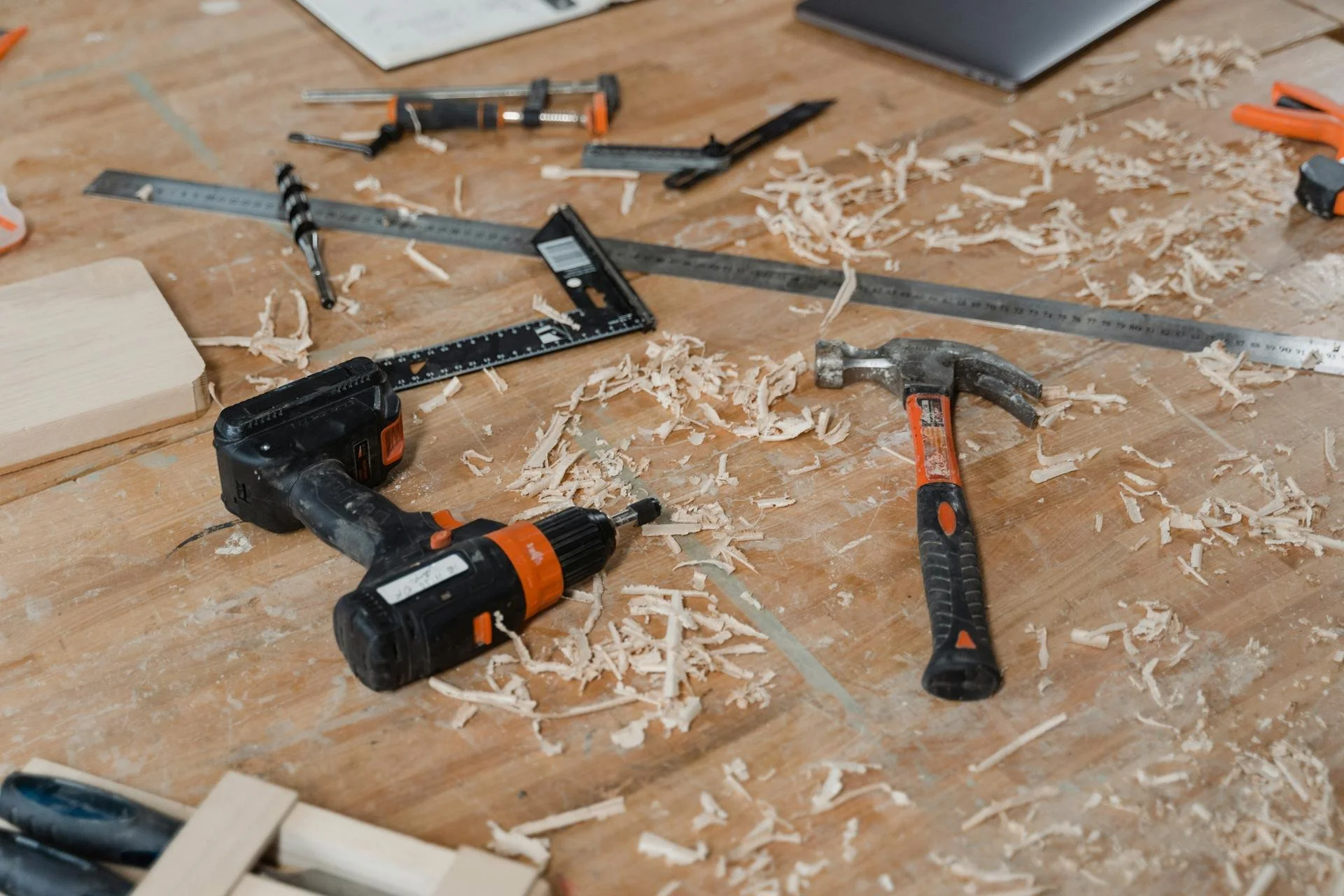 A cluttered woodworking workbench with tools including a cordless drill, hammer, square, and saw, surrounded by wood shavings and small pieces of cut wood.