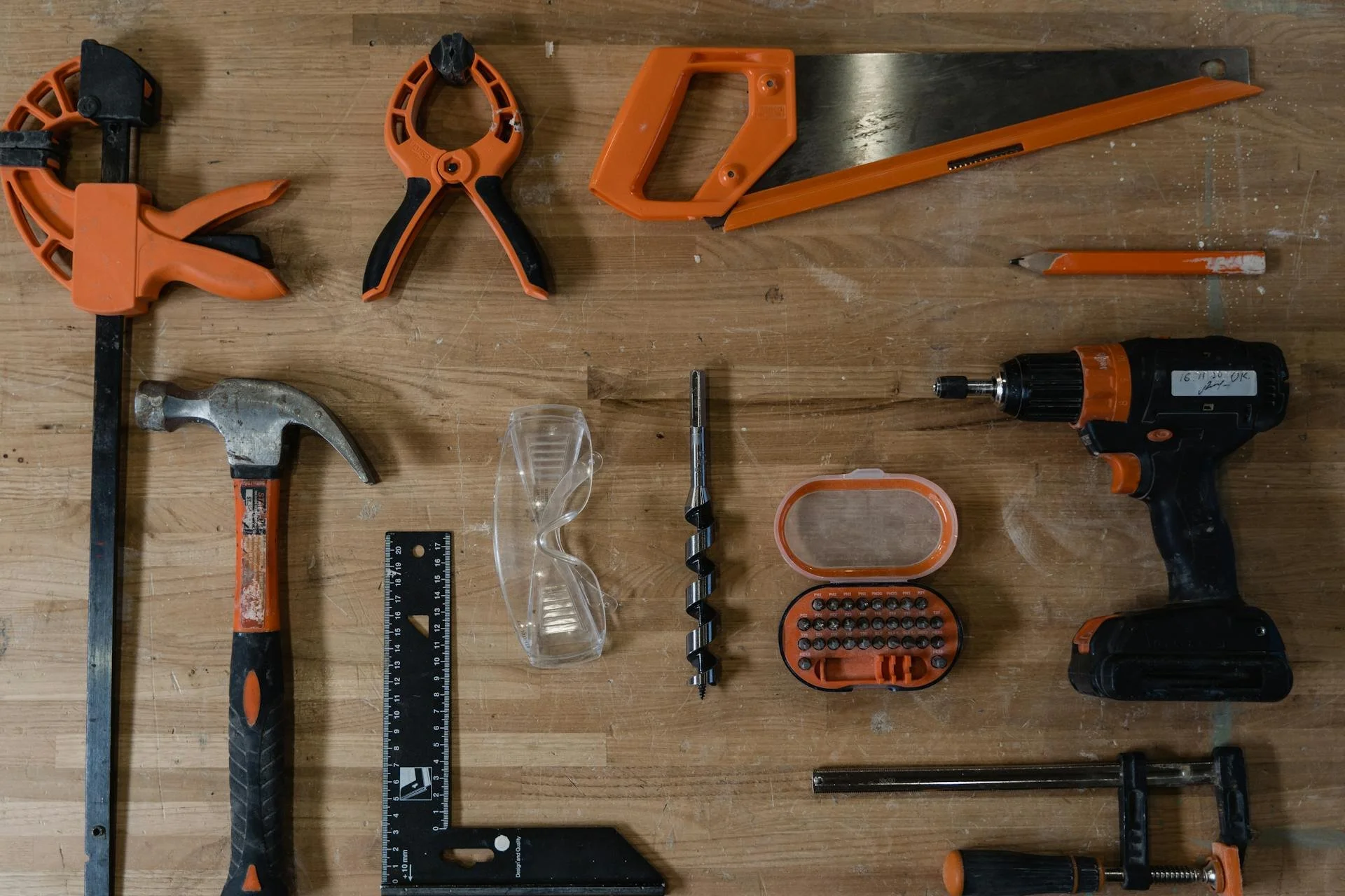 A wooden workspace with various tools including a hacksaw, adjustable wrench, cordless drill, drill bit, safety goggles, ruler, screwdriver bit, and screw organizer, mostly in orange and black colors.
