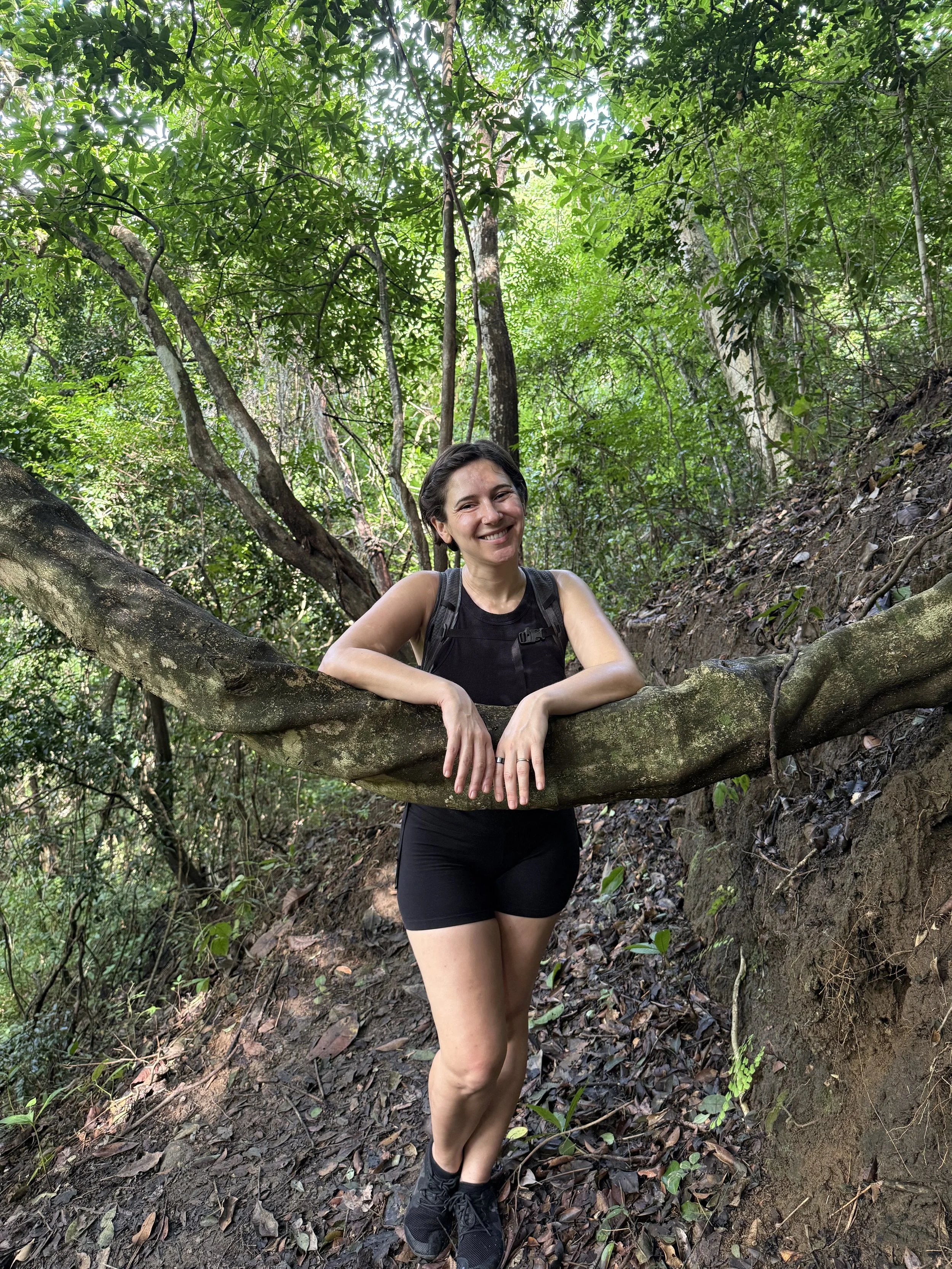 A woman with short, dark hair smiles as she leans on a large tree branch in a dense, green forest. She is wearing a black tank top, black shorts, and black athletic shoes, with a backpack on her shoulders. The ground is covered with leaf litter and small plants.