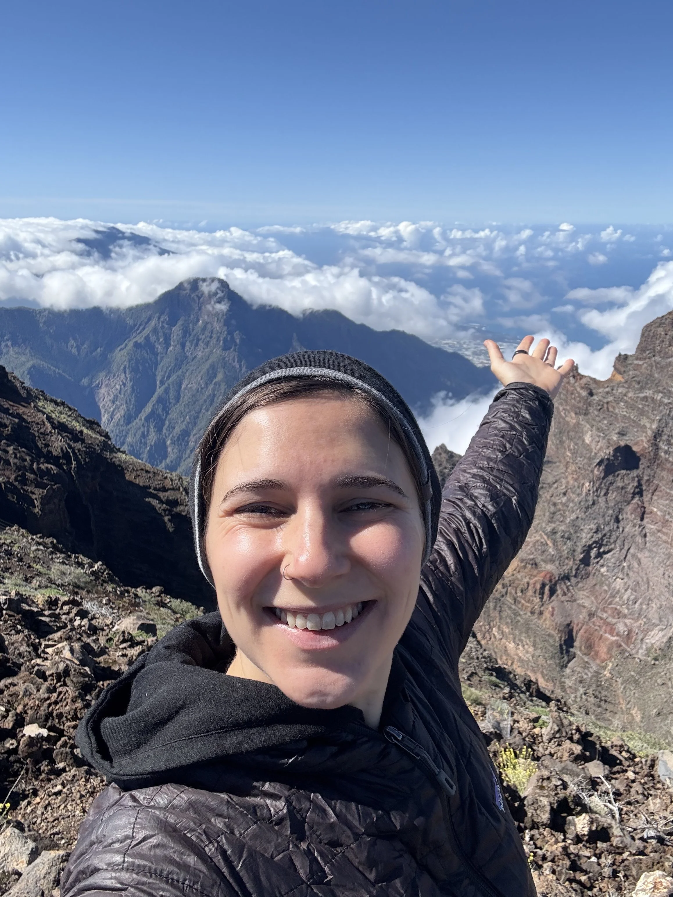 A woman taking a selfie on a mountain with a view of clouds and mountain peaks in the background. She is smiling and wearing a dark jacket and a beanie hat.