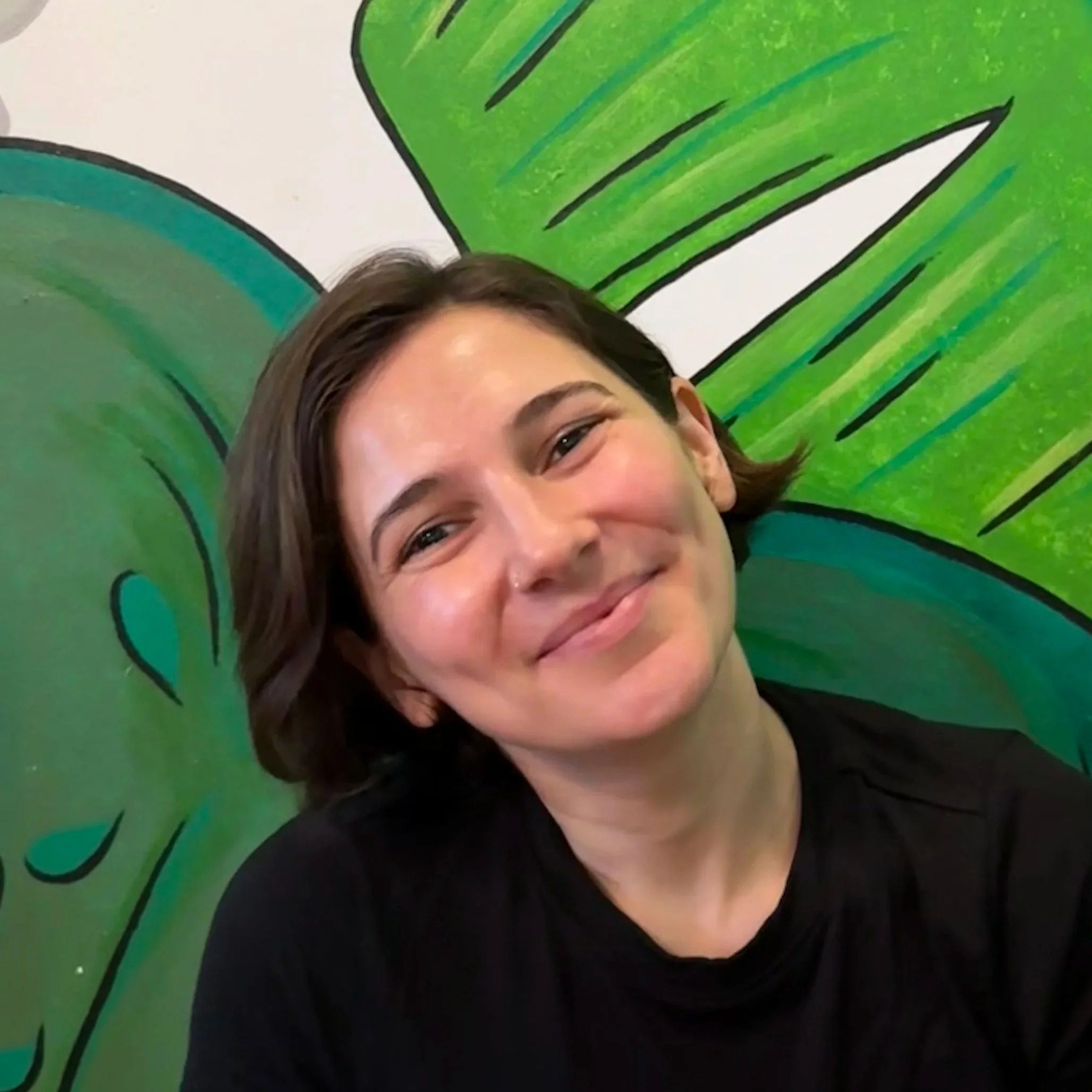 Close-up of a woman's face smiling, with a large green leaf painted on the wall behind her.