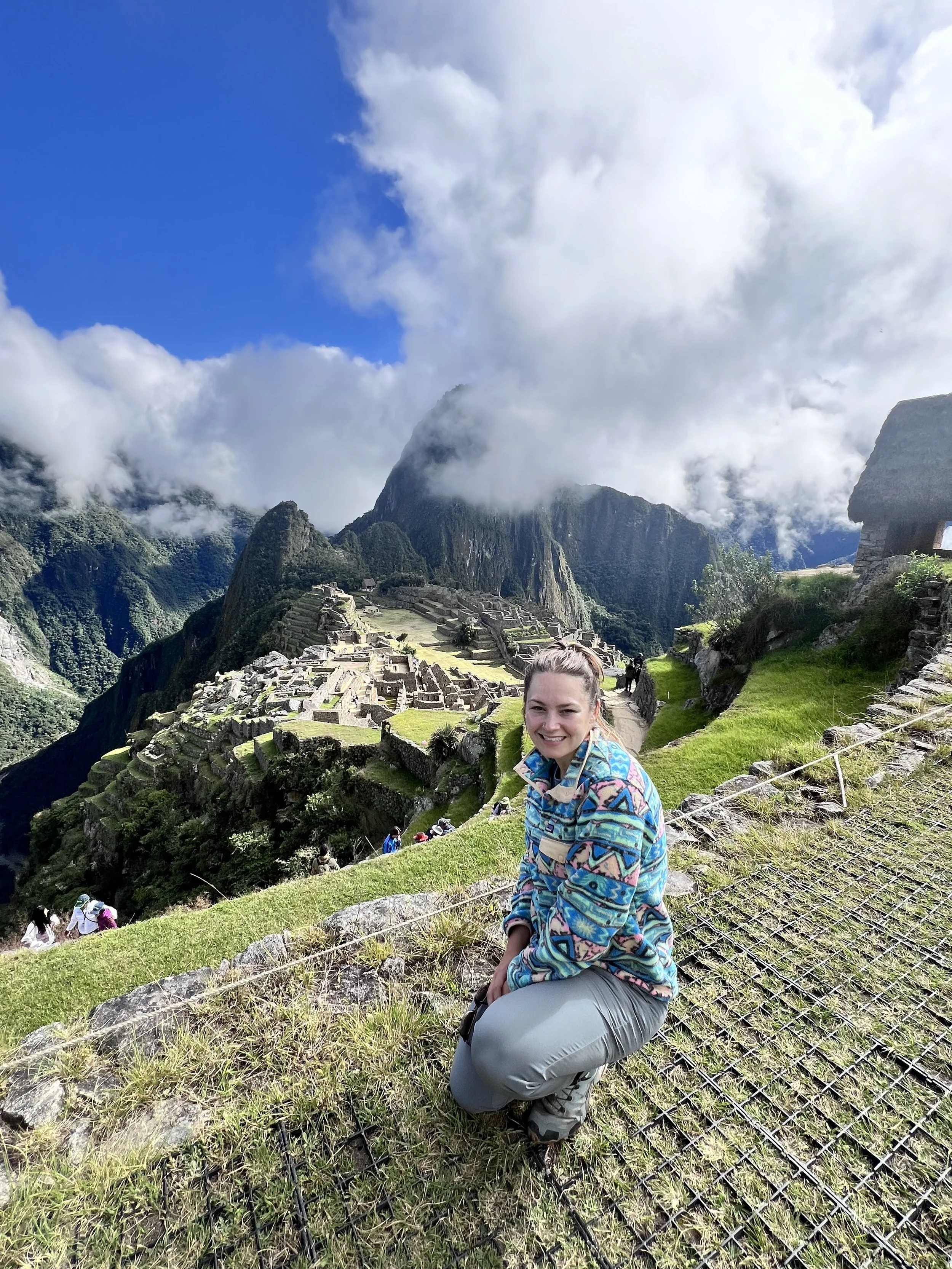 Young woman with blonde hair in a bun smiling at Machu Picchu with terraces, stone structures, and mountains in the background, during daytime with clouds and blue sky.
