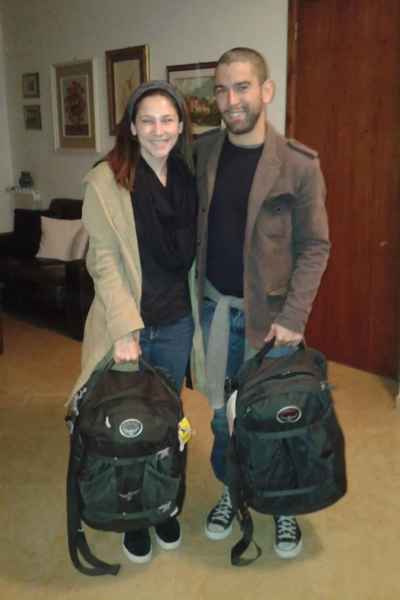 Two smiling people, a woman and a man, standing indoors with suitcases in front of them. The woman is on the left, wearing a beige jacket, black scarf, and sneakers. The man is on the right, wearing a brown jacket, dark shirt, and sneakers. They are in a room with framed pictures on the wall behind them.