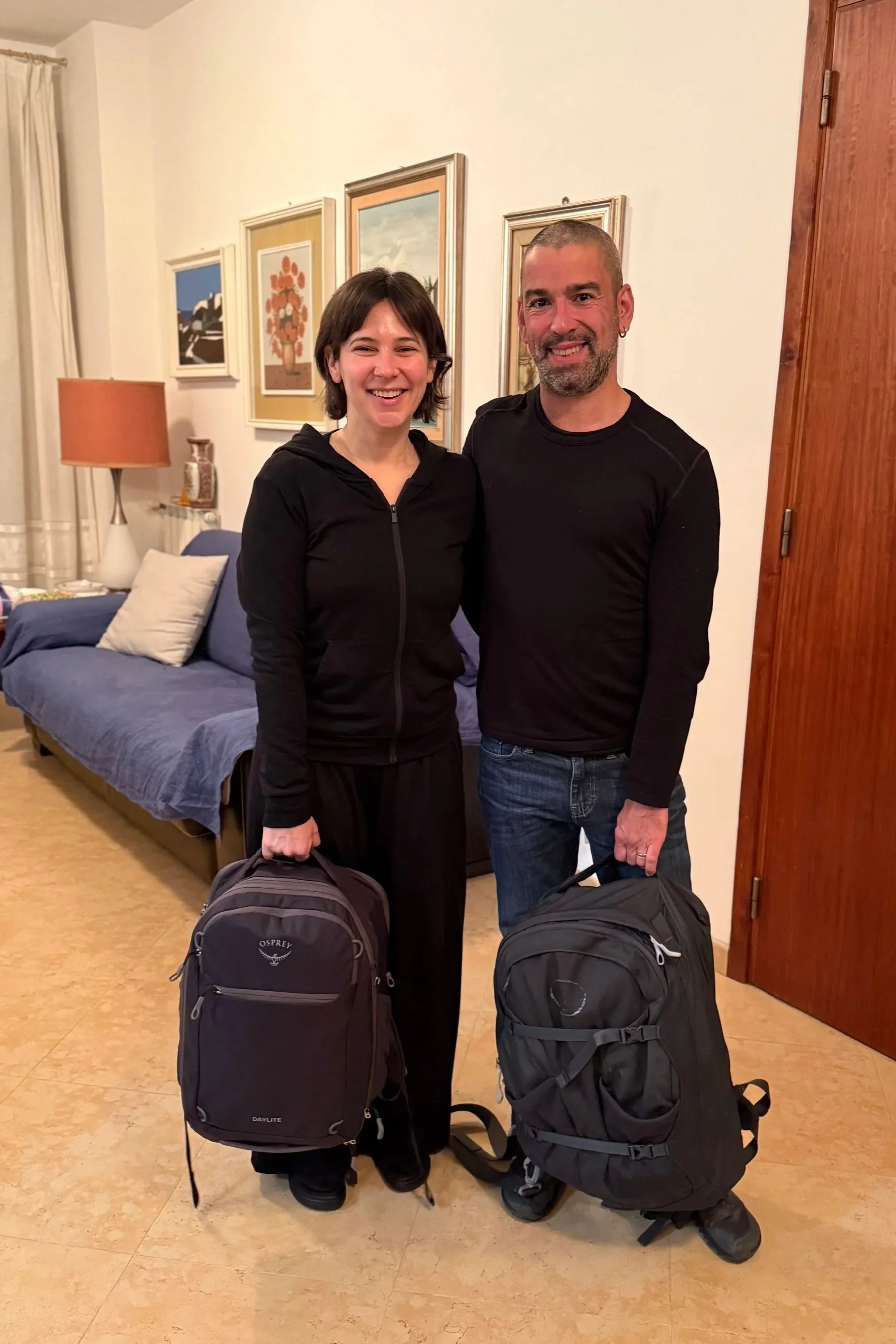 A woman and a man standing inside a room, both smiling, holding backpacks. The room has a blue couch with pillows, framed artwork on the wall, a wooden door, and a lamp on a side table.