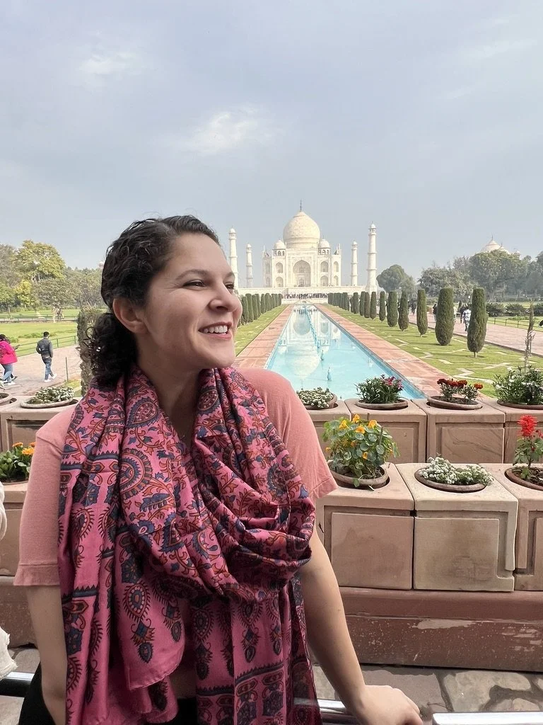 A woman smiling in front of the Taj Mahal with a pink and purple patterned scarf, on a cloudy day.