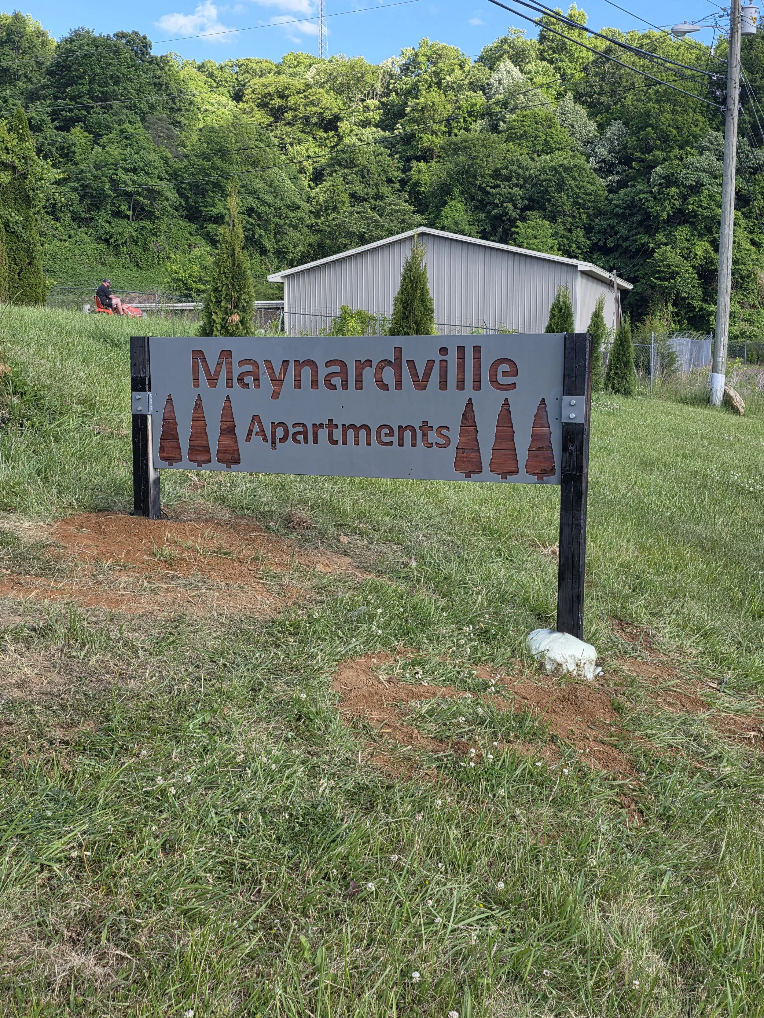 Sign that reads 'Maynardville Apartments' with illustrations of pine trees. Background shows a grassy area, a gray building, and a lush green hillside.