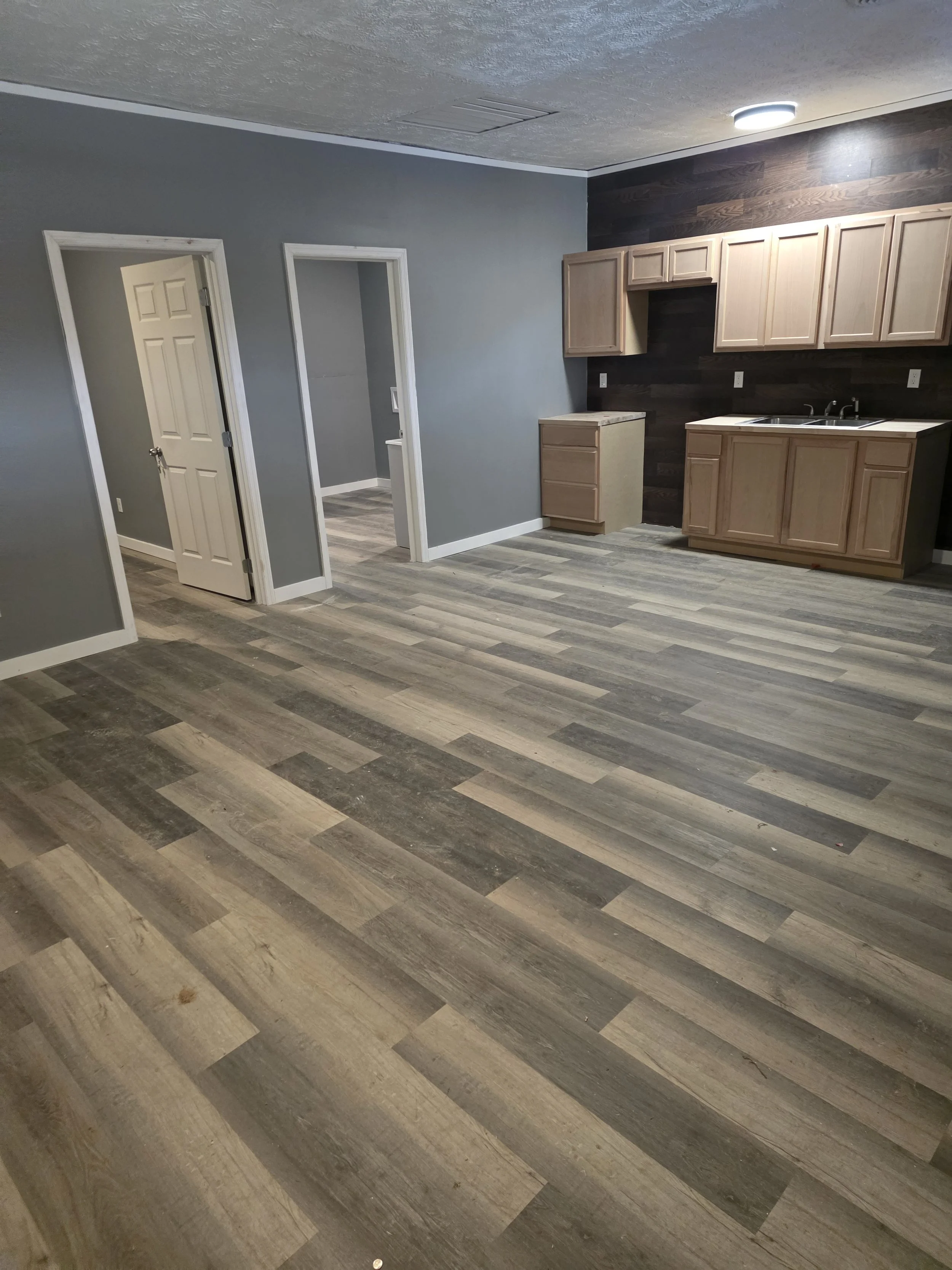 An empty kitchen with wood cabinets, a sink, gray wall, wood-look flooring, and a ceiling light.
