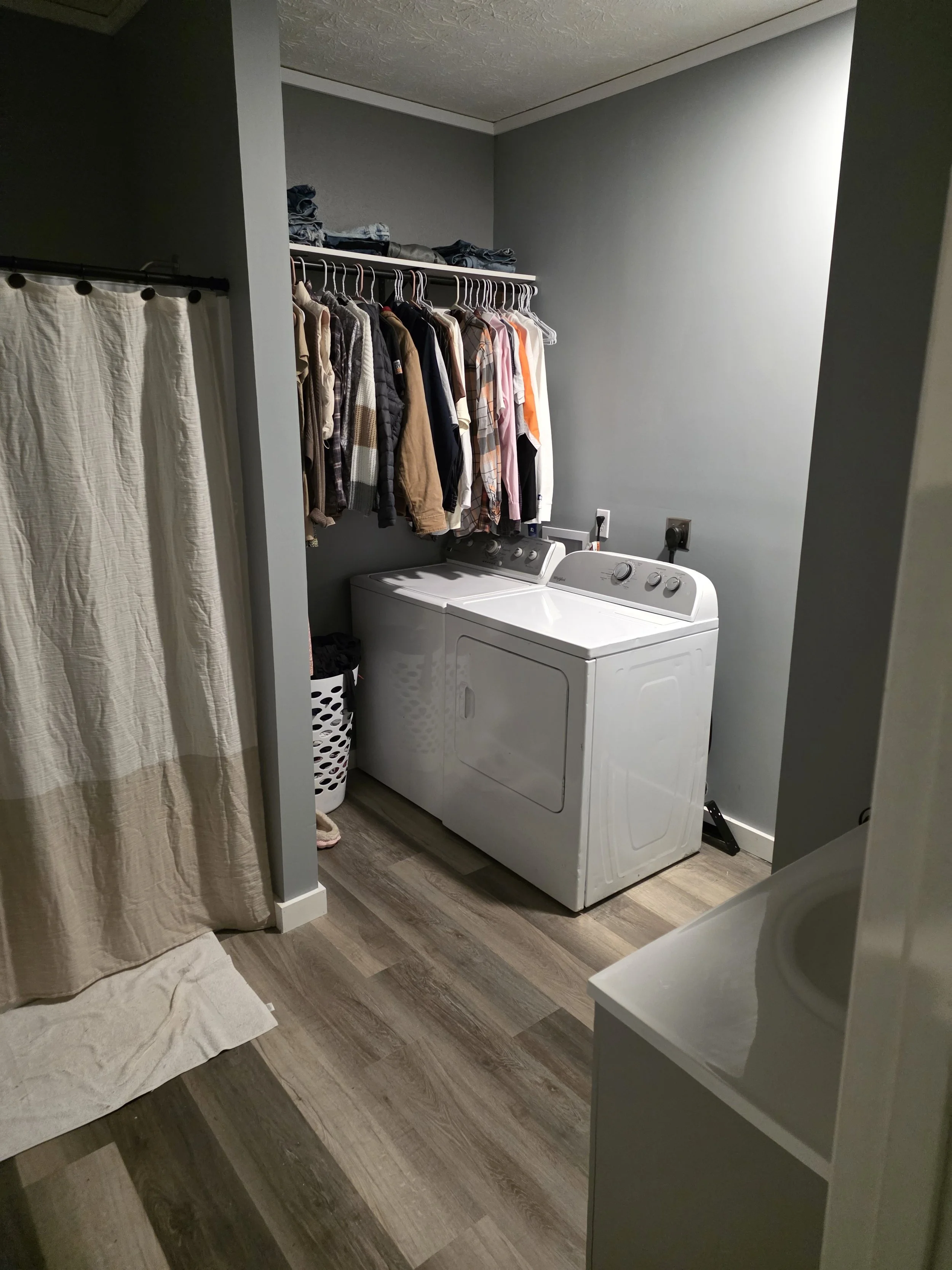 A laundry room with a washing machine, a hanging rack with clothes, and a laundry basket. The room has gray walls and wood-style flooring.