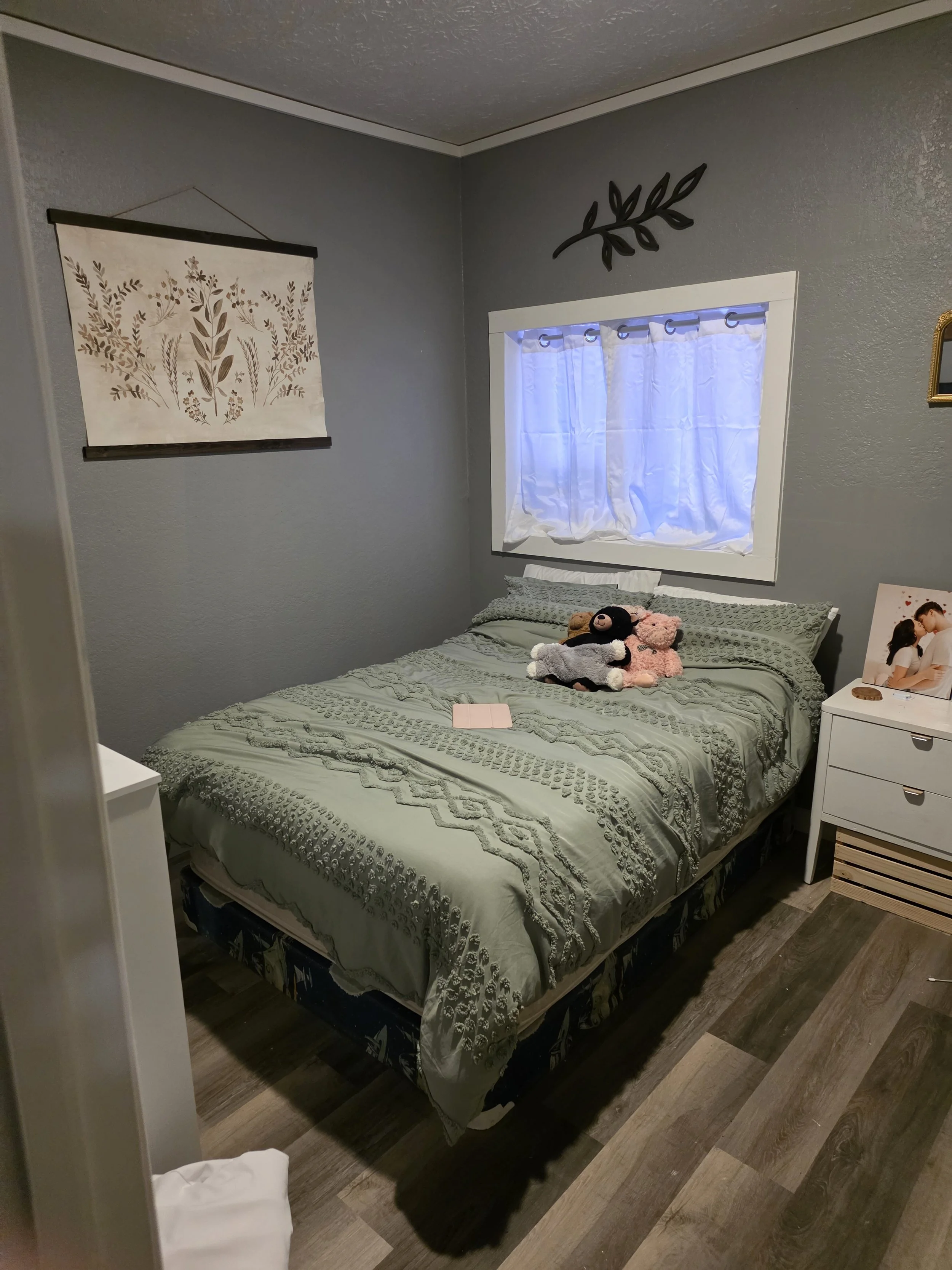 A bedroom with gray walls, a bed with green bedding, and stuffed animals on it. There is a small white nightstand with a framed photo and a wicker basket. A window with white curtains is above the bed, and wall decor includes a black leaf branch above the window and a botanical print on the opposite wall. The room has wood-look flooring.