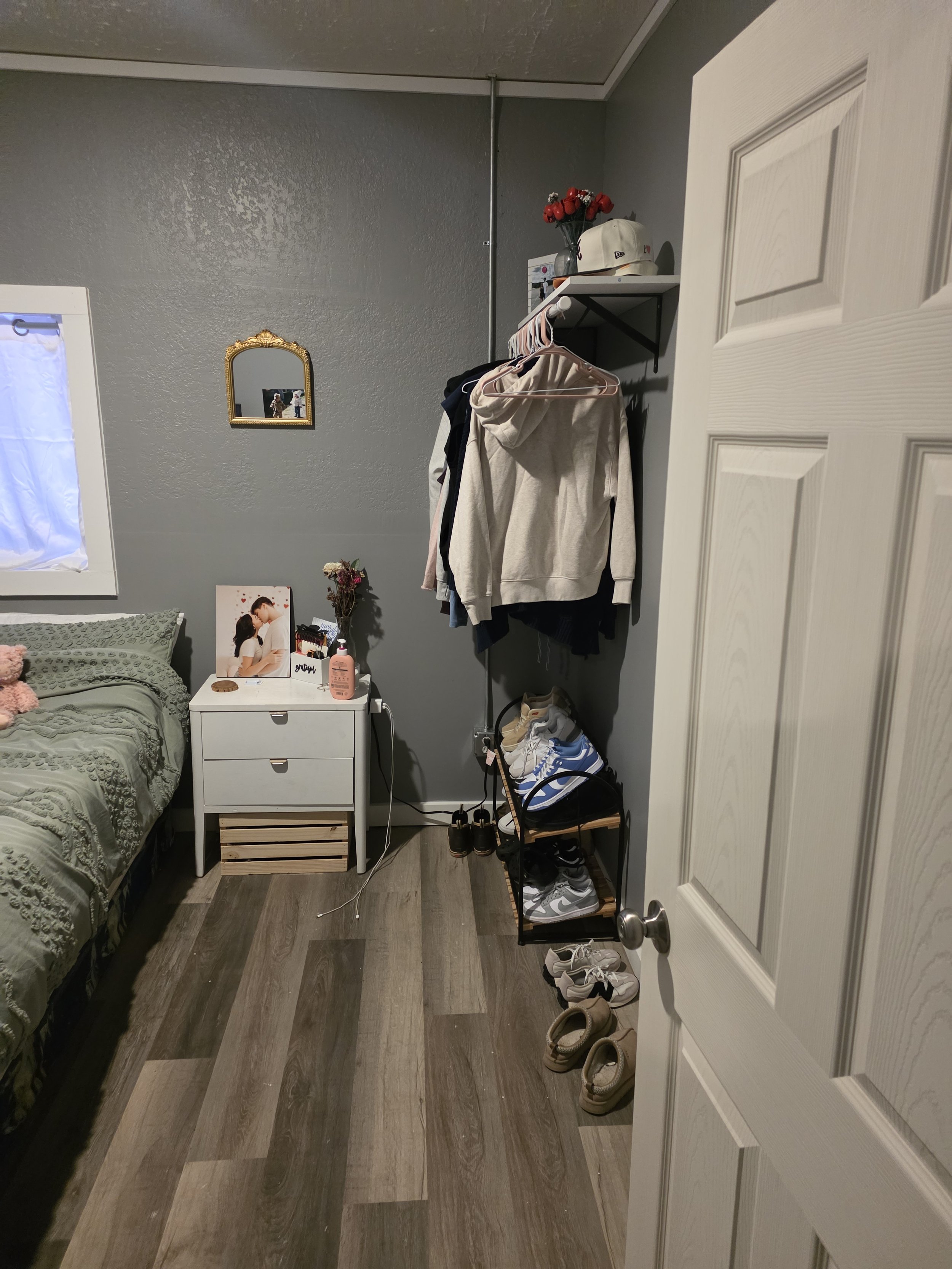 A bedroom corner with a bed, nightstand, wall mirror, clothing rack, shoes, and various decorative items.