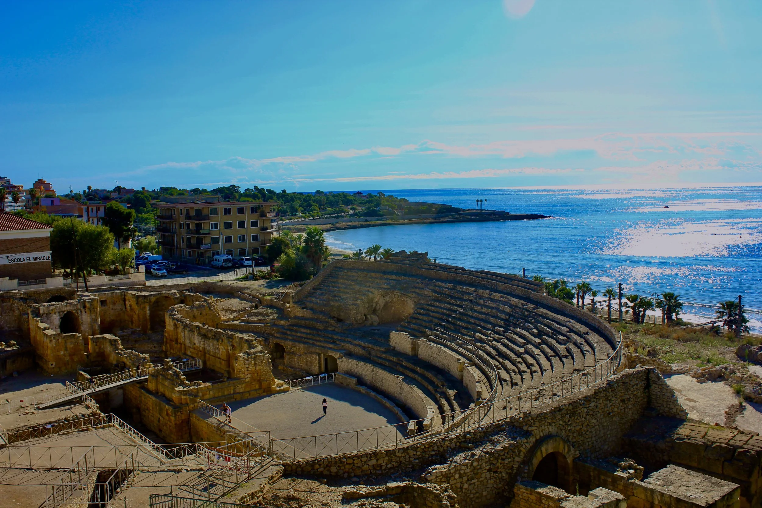 Ancient Roman amphitheater ruins overlooking the ocean with modern buildings and a beach in the background.