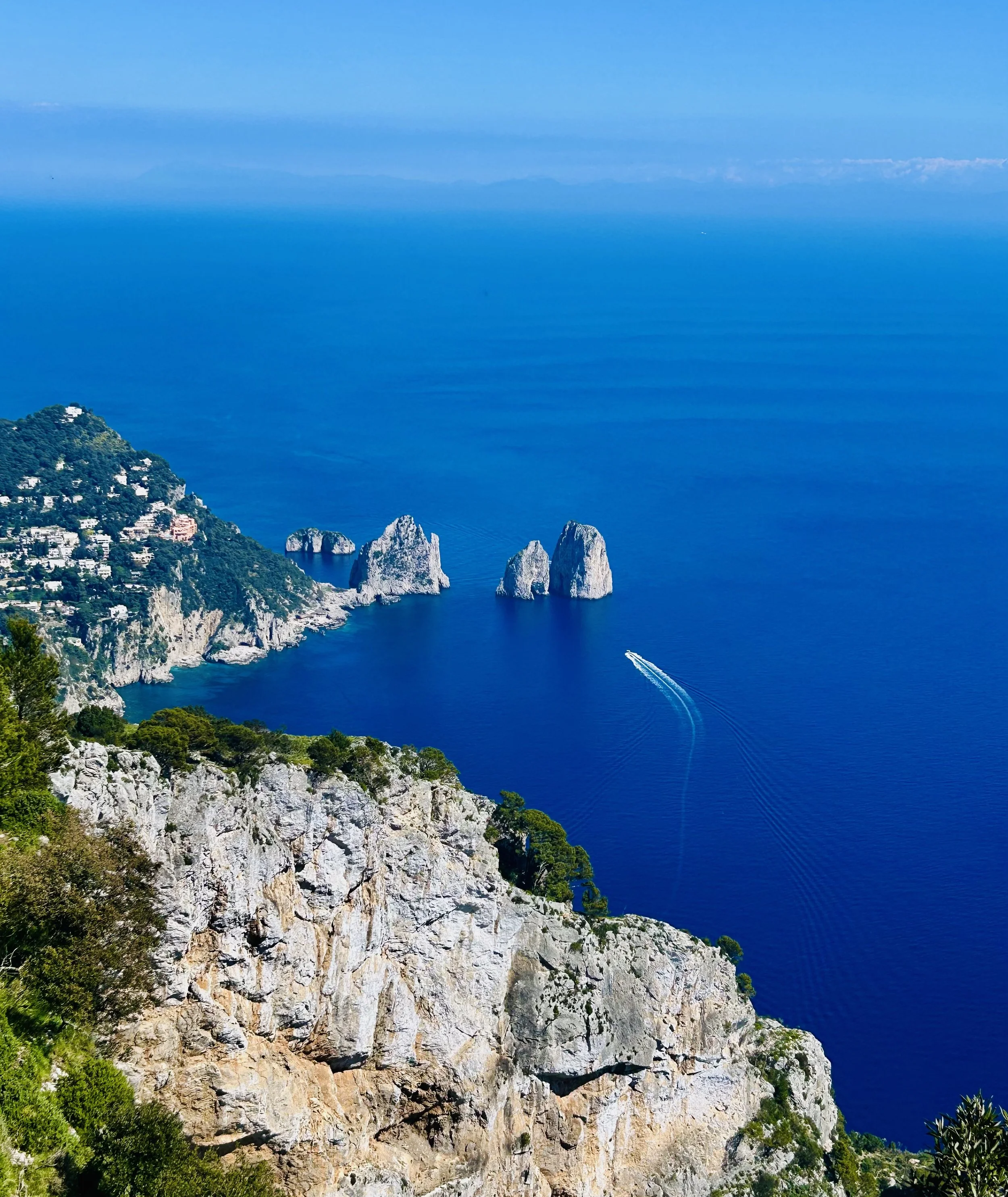 Cliffside view of a coastal area with rocky formations, green vegetation, and a calm blue sea stretching into the horizon under a clear sky.
