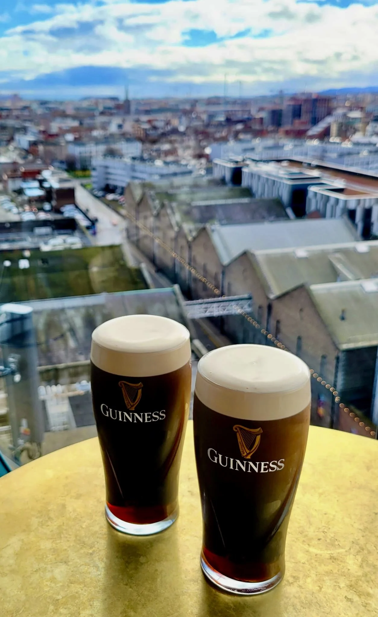Two pints of Guinness beer on a yellow table in front of a large window overlooking city buildings and a cloudy sky.