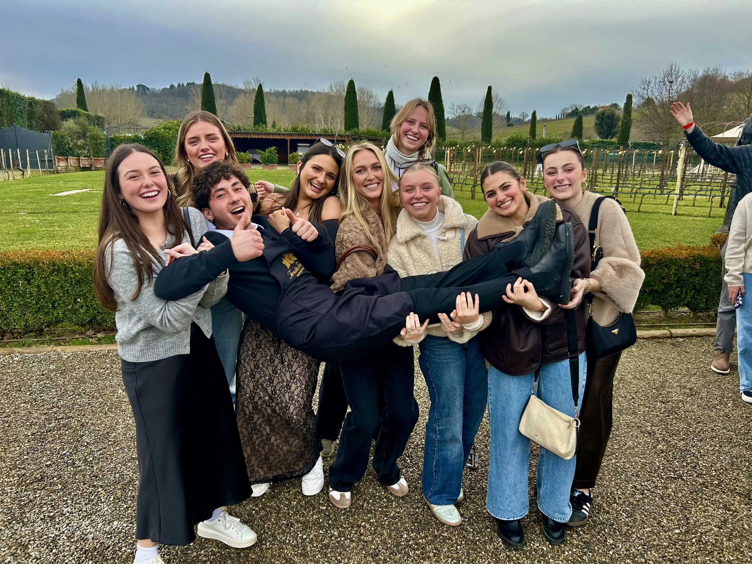 Group of ten young adults smiling and posing together outdoors in a vineyard or garden setting with trees and hills in the background on a cloudy day.