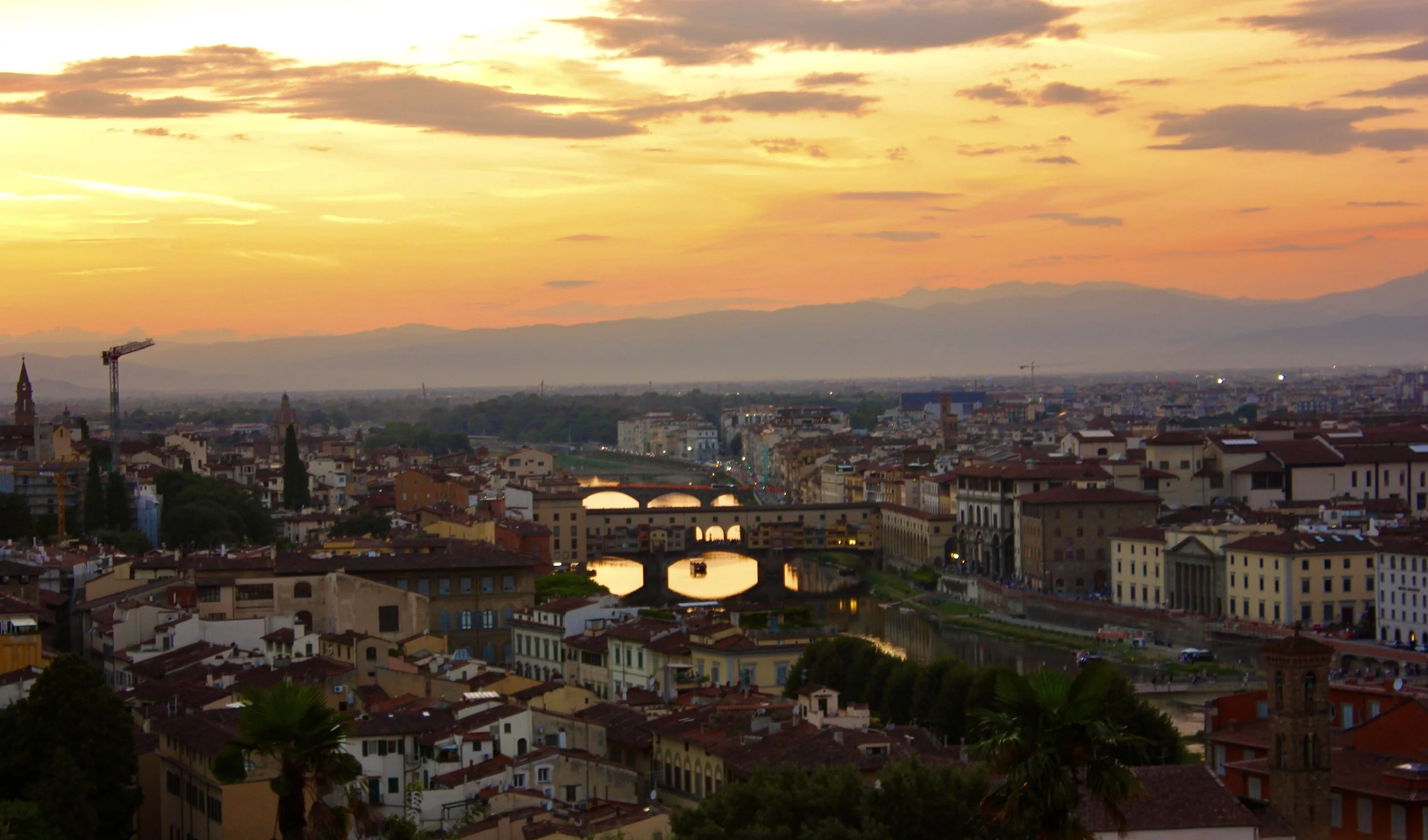 Sunset over a city with a river running through it, multiple bridges, and a mountainous background.
