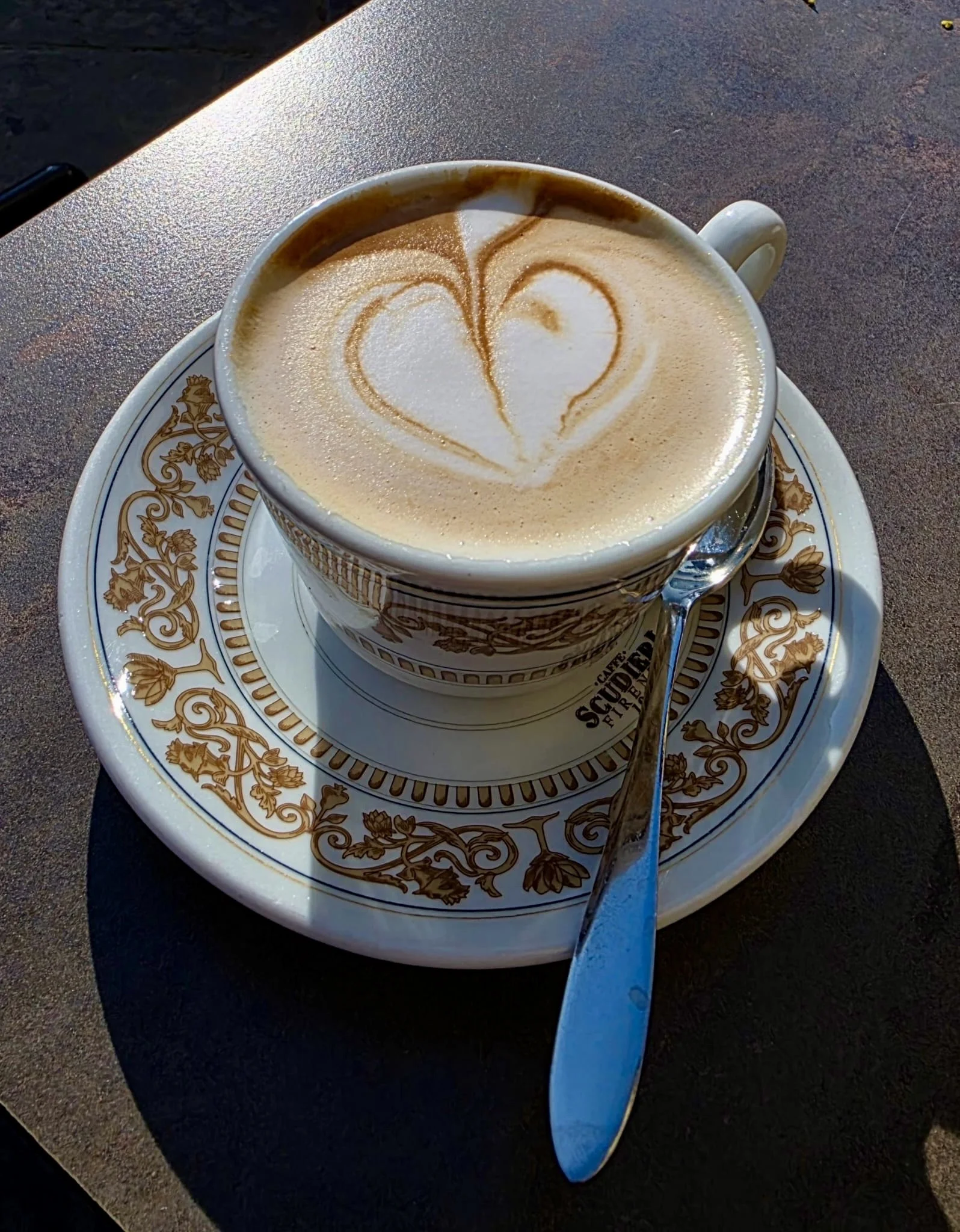A cup of frothy coffee with heart-shaped latte art on top, placed on a decorative saucer with floral patterns. A silver spoon rests on the saucer, and the setup is on a dark textured table.