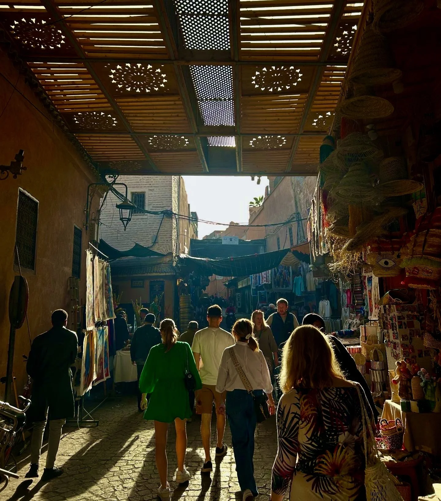 People walking through a bustling outdoor market with shops and stalls on both sides, sunlight casting long shadows, and a decorative wooden canopy overhead.