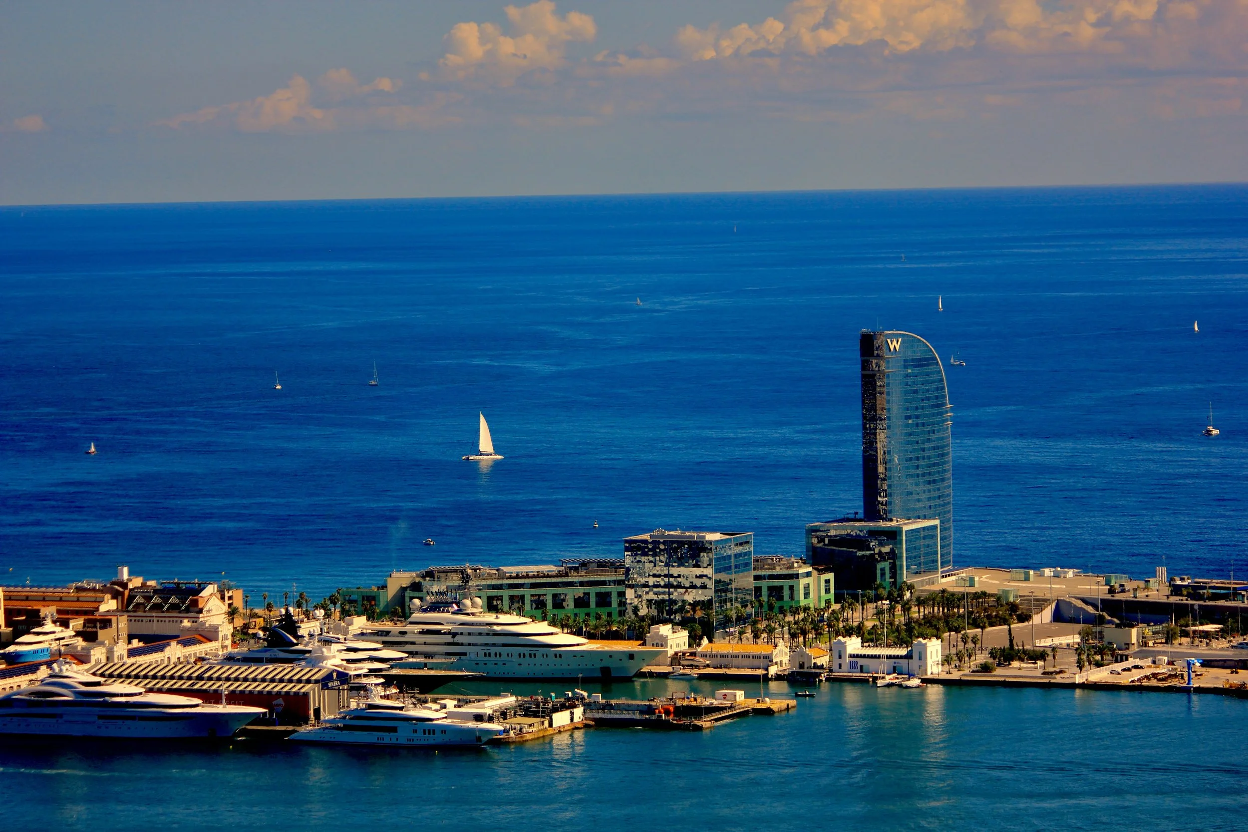City waterfront with yachts and sailboats, tall modern buildings, and a blue ocean under a partly cloudy sky.