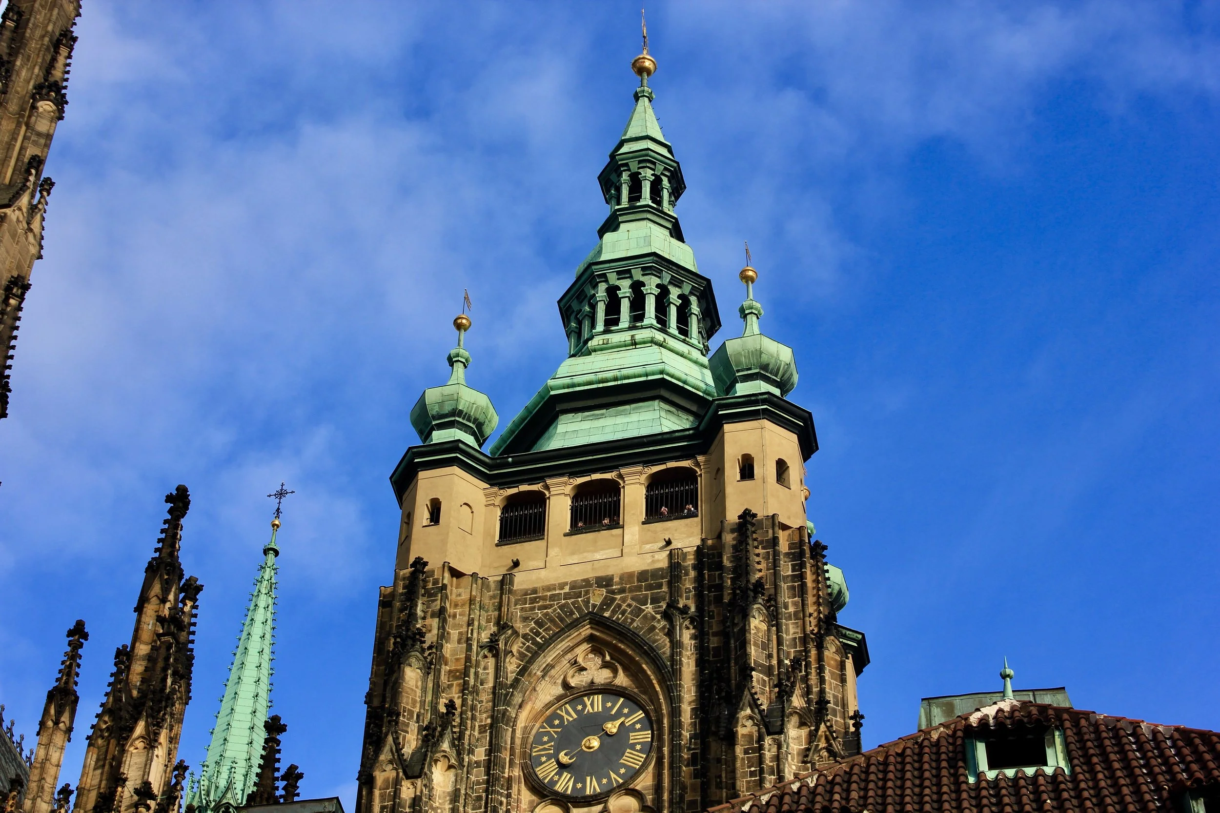 Close-up view of a historic church tower with a clock, tall spires, and intricate Gothic architectural details against a clear blue sky.