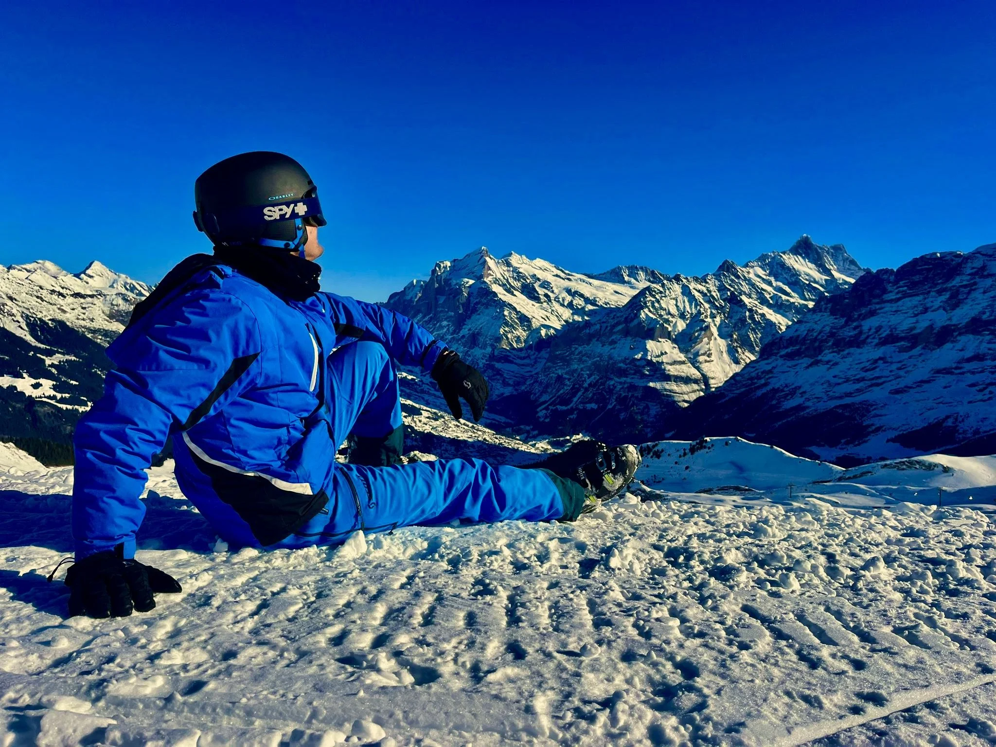 A person in a blue jacket, black helmet, and snow gear sitting on snow-covered ground with snow-capped mountains in the background under a clear blue sky.