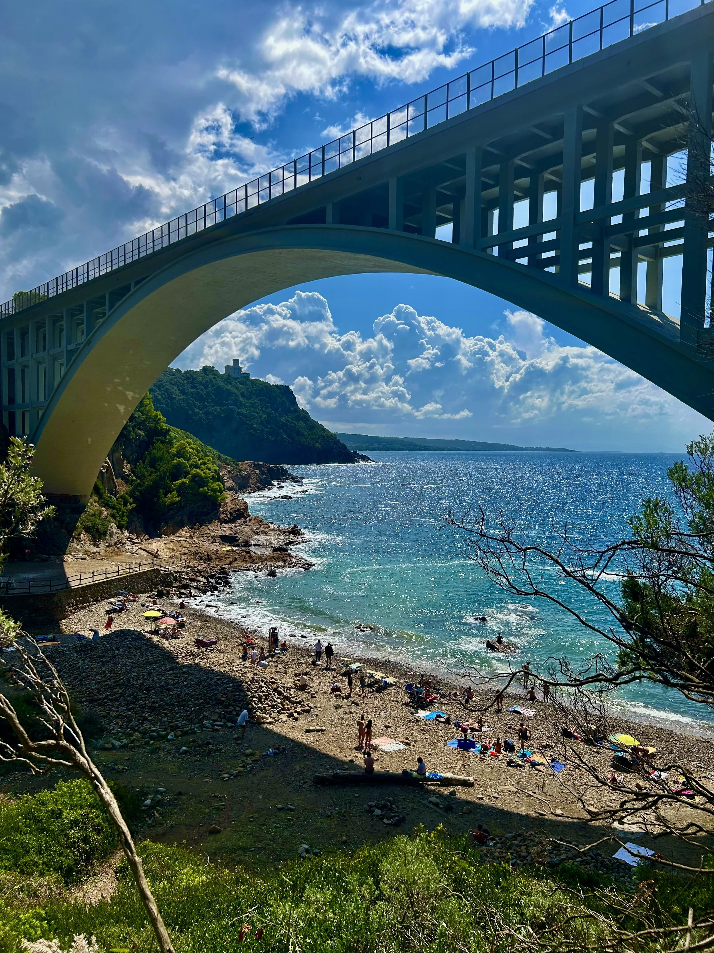 A scenic view of a bridge over a coastal area with a beach. The sky is partly cloudy with sunlight shining through. People are relaxing on the beach with umbrellas, and there is lush greenery in the foreground and a rocky hill in the background.