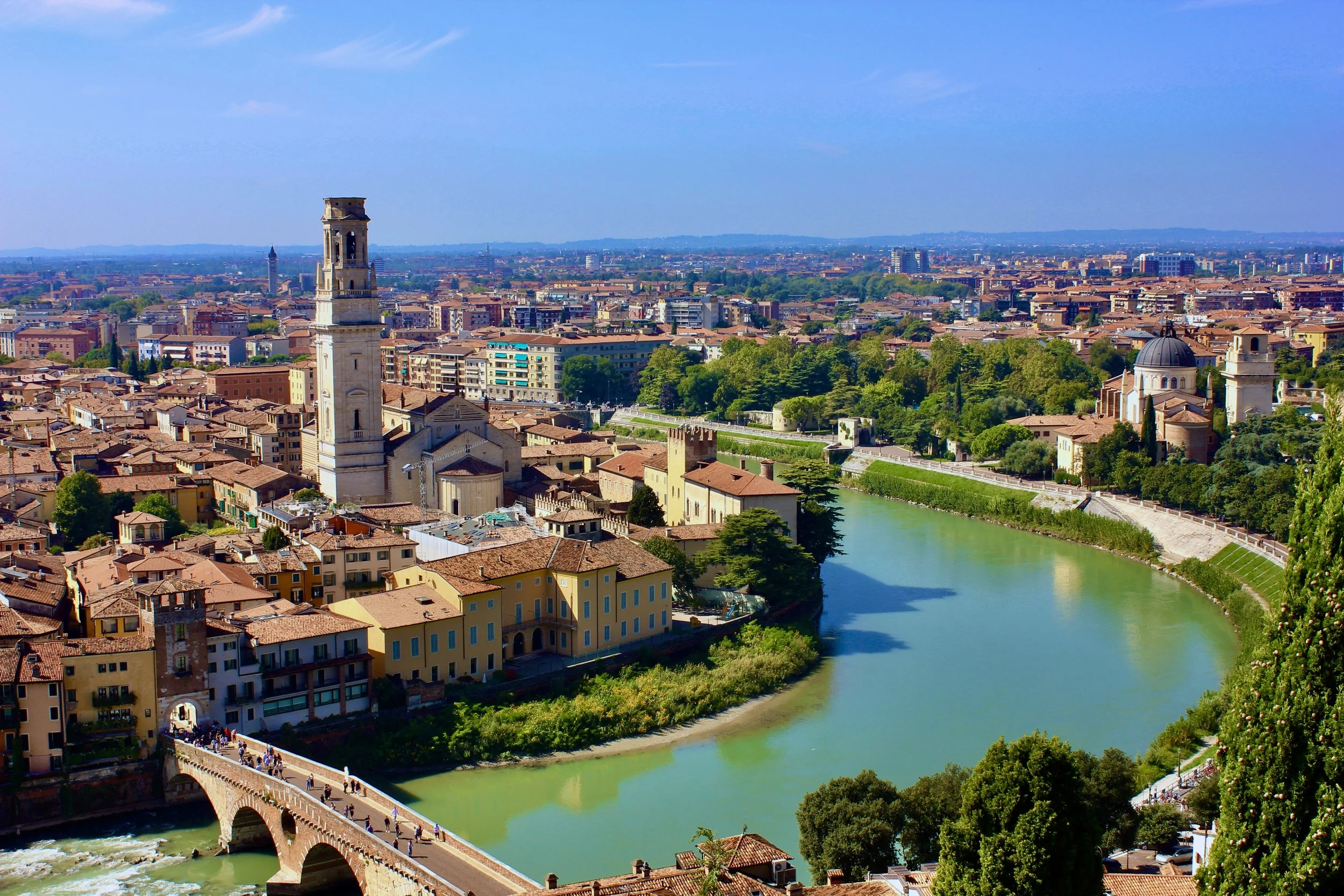 A view of a city with historic buildings, a bridge over a river, and a prominent tall clock tower against a clear blue sky.