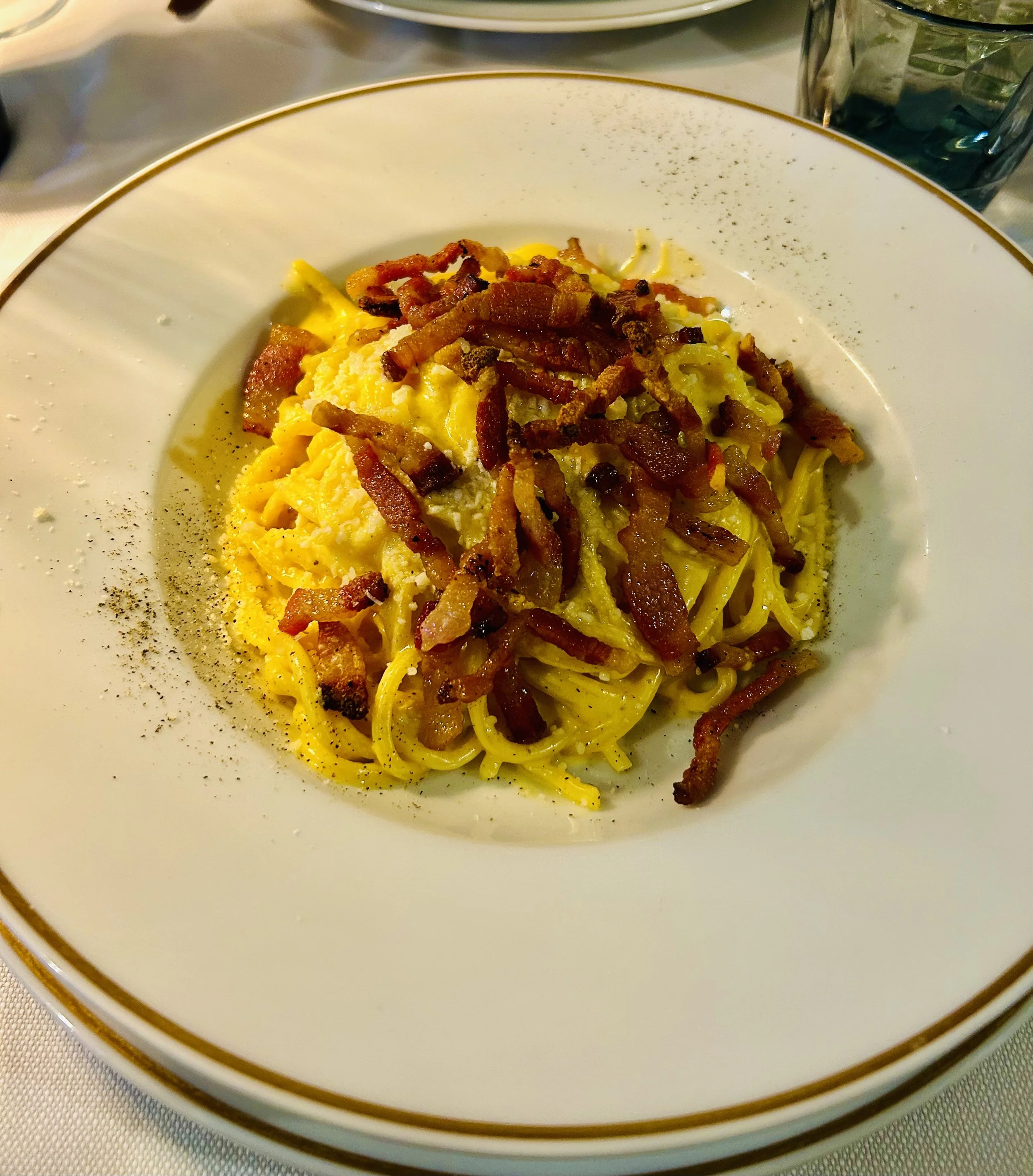 Close-up of a white plate with a gold rim containing scrambled eggs topped with crispy bacon strips, garnished with black pepper.