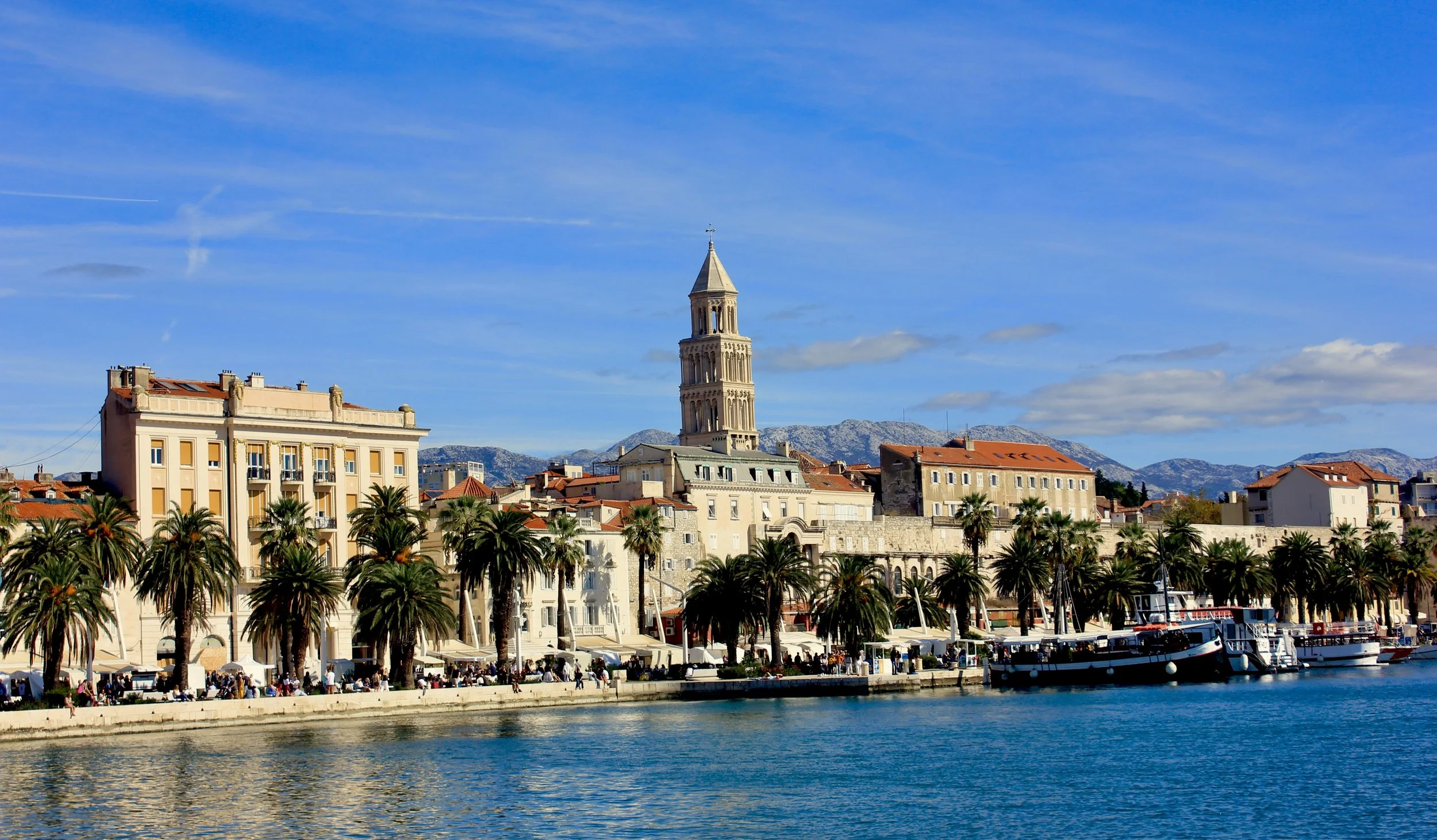 Cityscape with historic tower, Mediterranean-style buildings with orange roofs, palm trees along the waterfront, boats docked in the harbor, mountains in the background, and a partly cloudy sky.