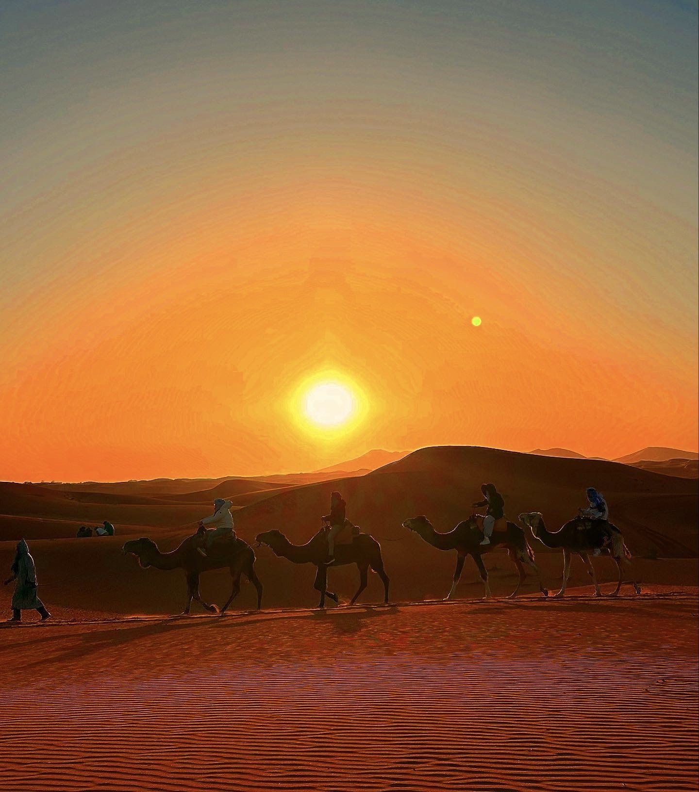Silhouettes of people riding camels across desert dunes at sunset, with a bright sun and a small greenish celestial body in the sky.