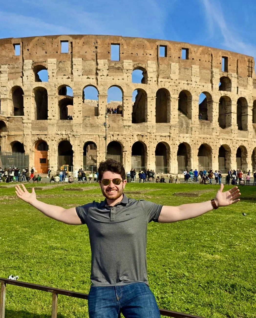 A man with sunglasses smiling and spreading his arms wide in front of the Colosseum in Rome, Italy, under a blue sky.