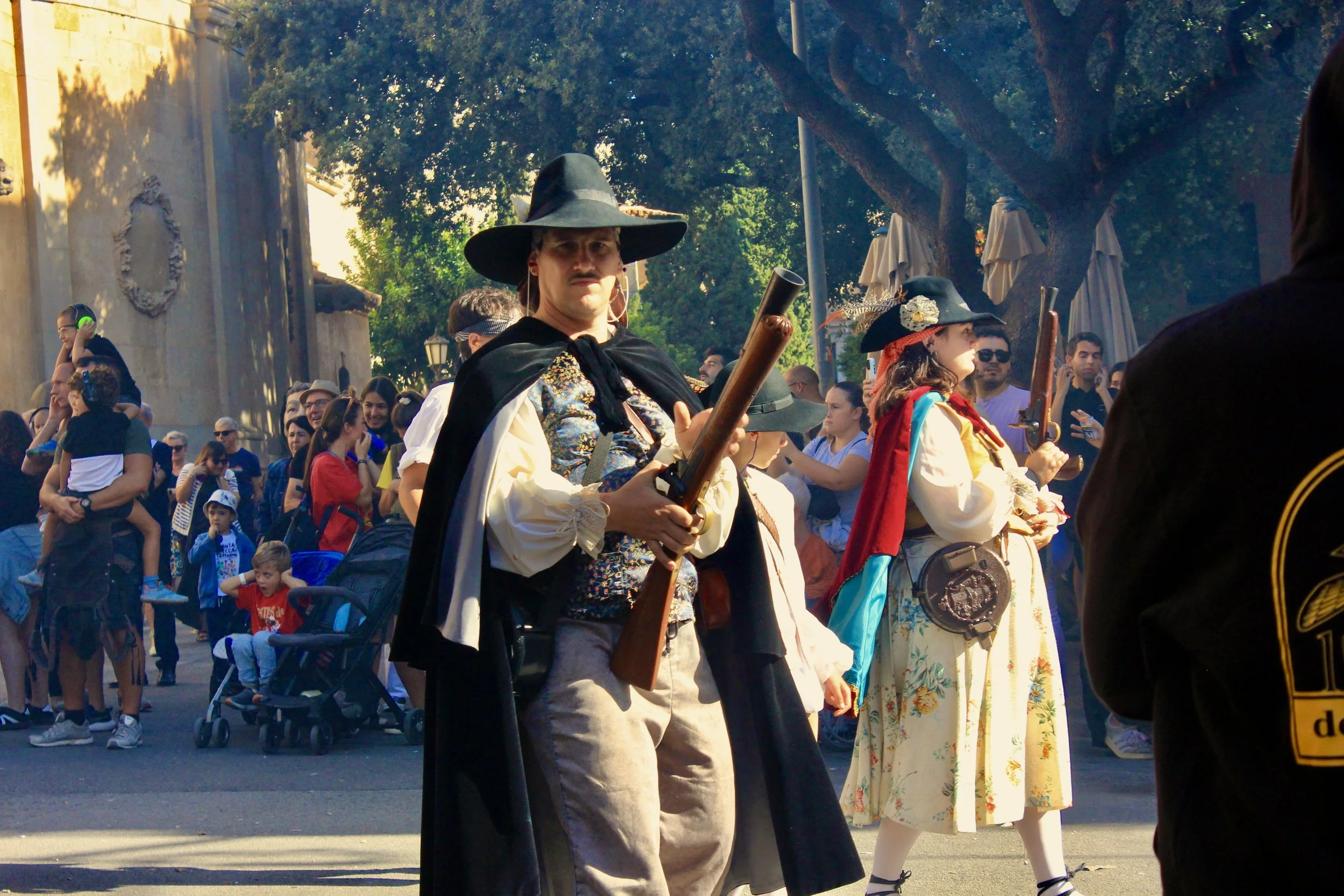 People dressed in historical costumes participating in a public event or parade outdoors, with trees and a crowd in the background.