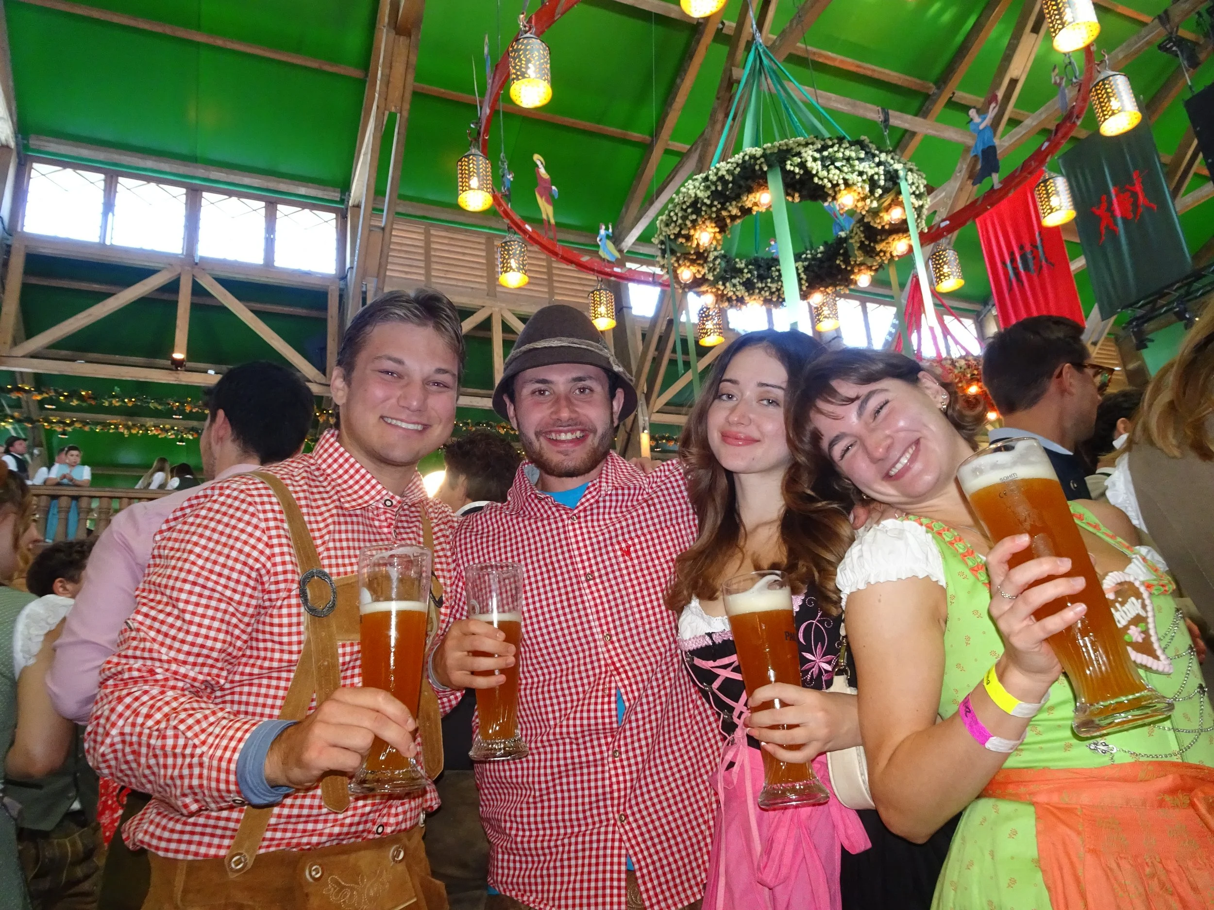 People in traditional German clothing celebrating at an indoor event, holding large beer mugs, with festive decorations and a green ceiling in the background.