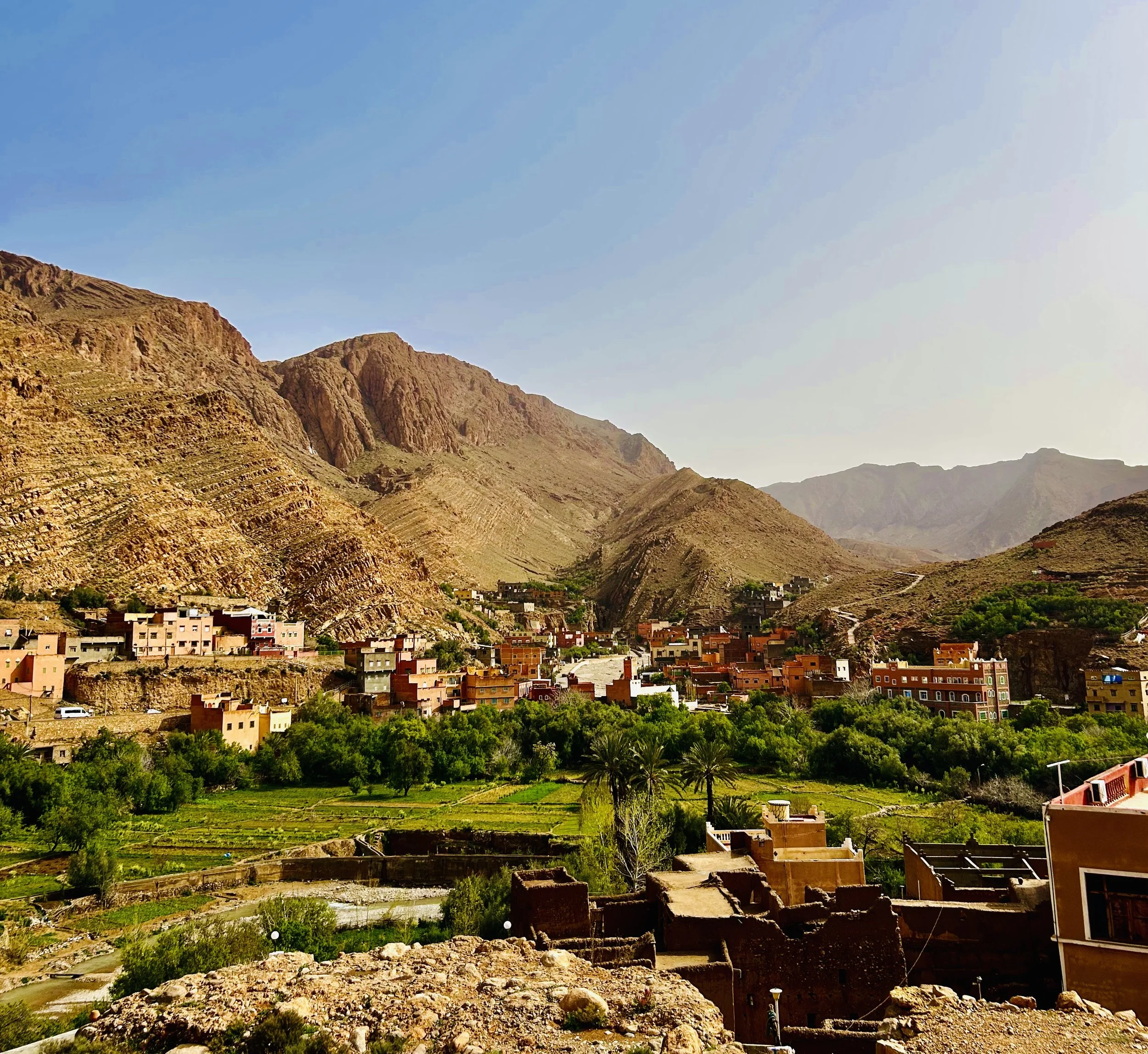 A small village with colorful houses surrounded by green trees, with large rocky mountains in the background under a clear blue sky.